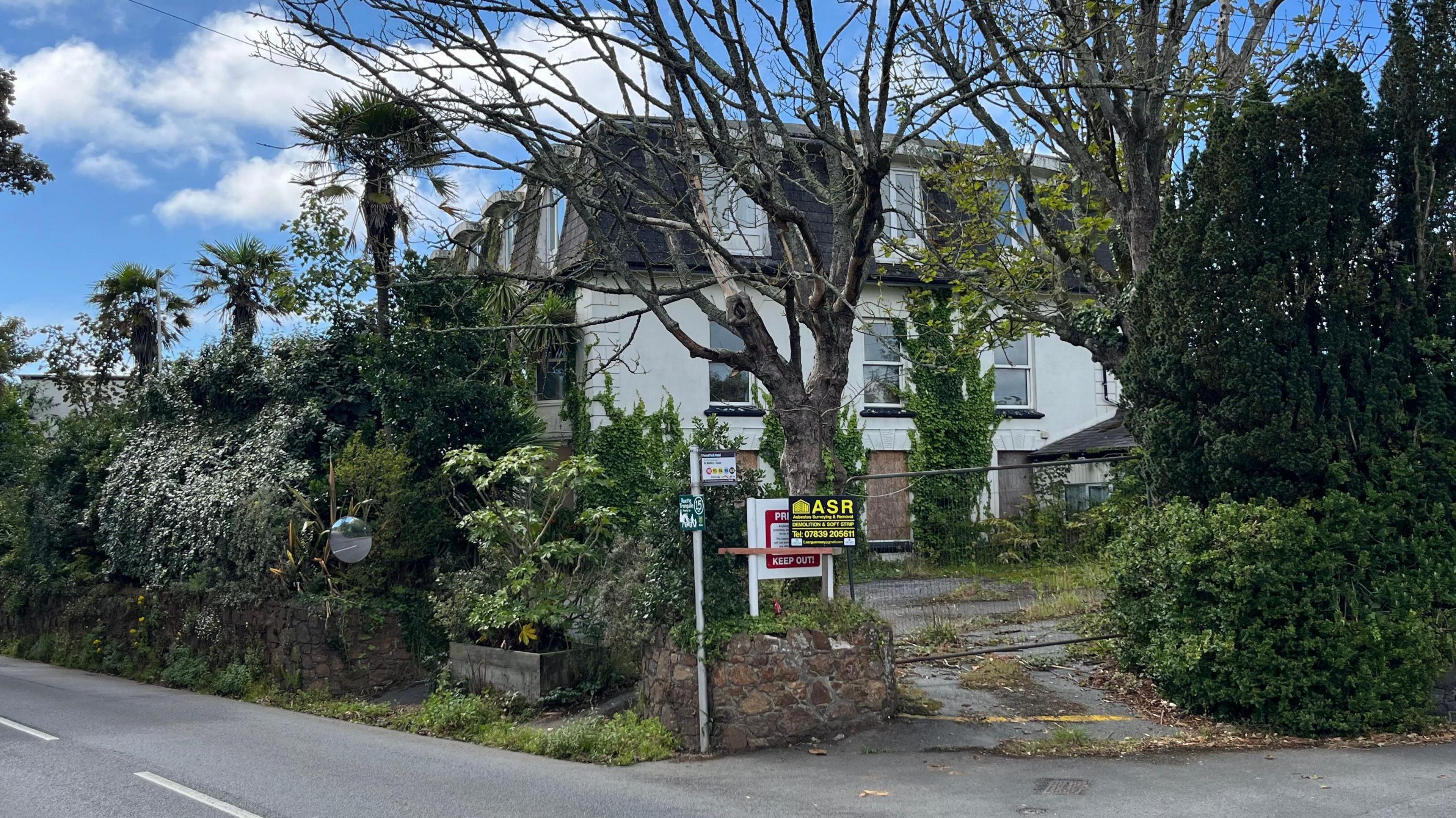 The St Margaret's Lodge, a hotel that has become derelict, behind a group of trees. There is a black and yellow sign for asbestos removal.