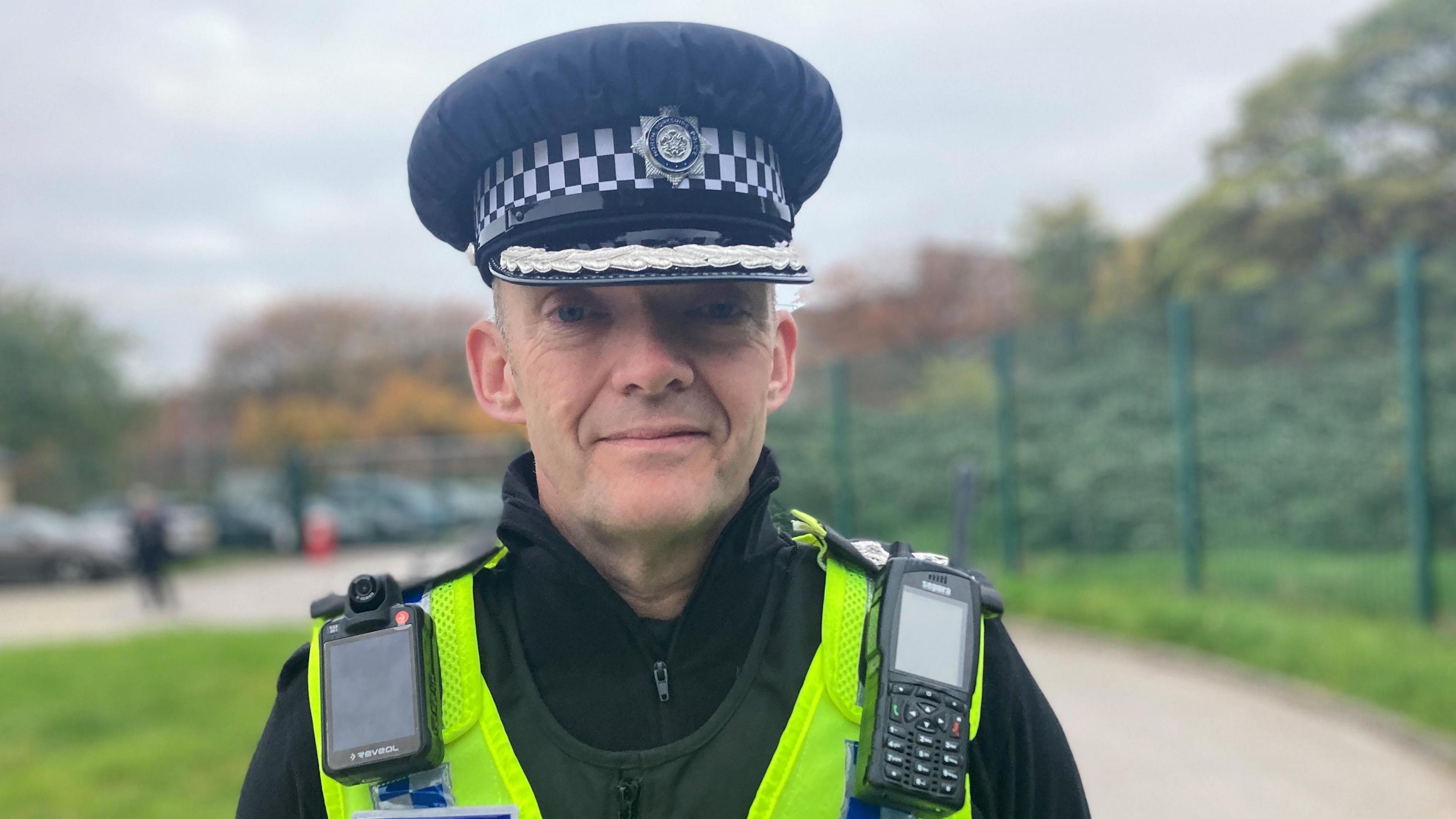 Deputy Chief Constable Scott Bisset of North Yorkshire Police looks at the camera. He is wearing a peaked uniform hat in black, a black polo style top and body protective gear and a yellow vest over all with a body camera and radio