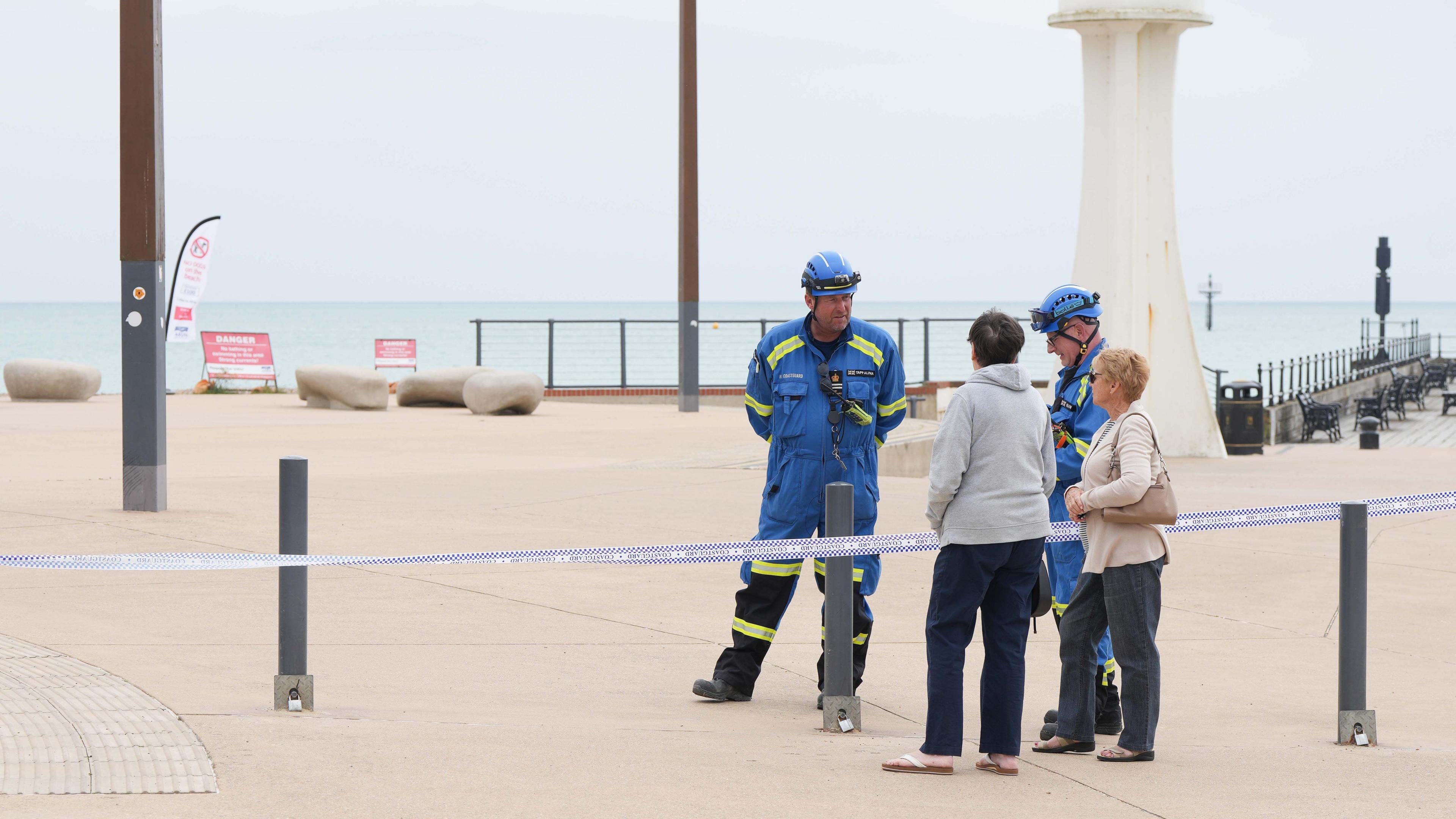 Two men in blue overalls and hard hats stood on a promenade. They are talking to two women on the other side of a blue tape cordon and the promenade is otherwise clear of people.
