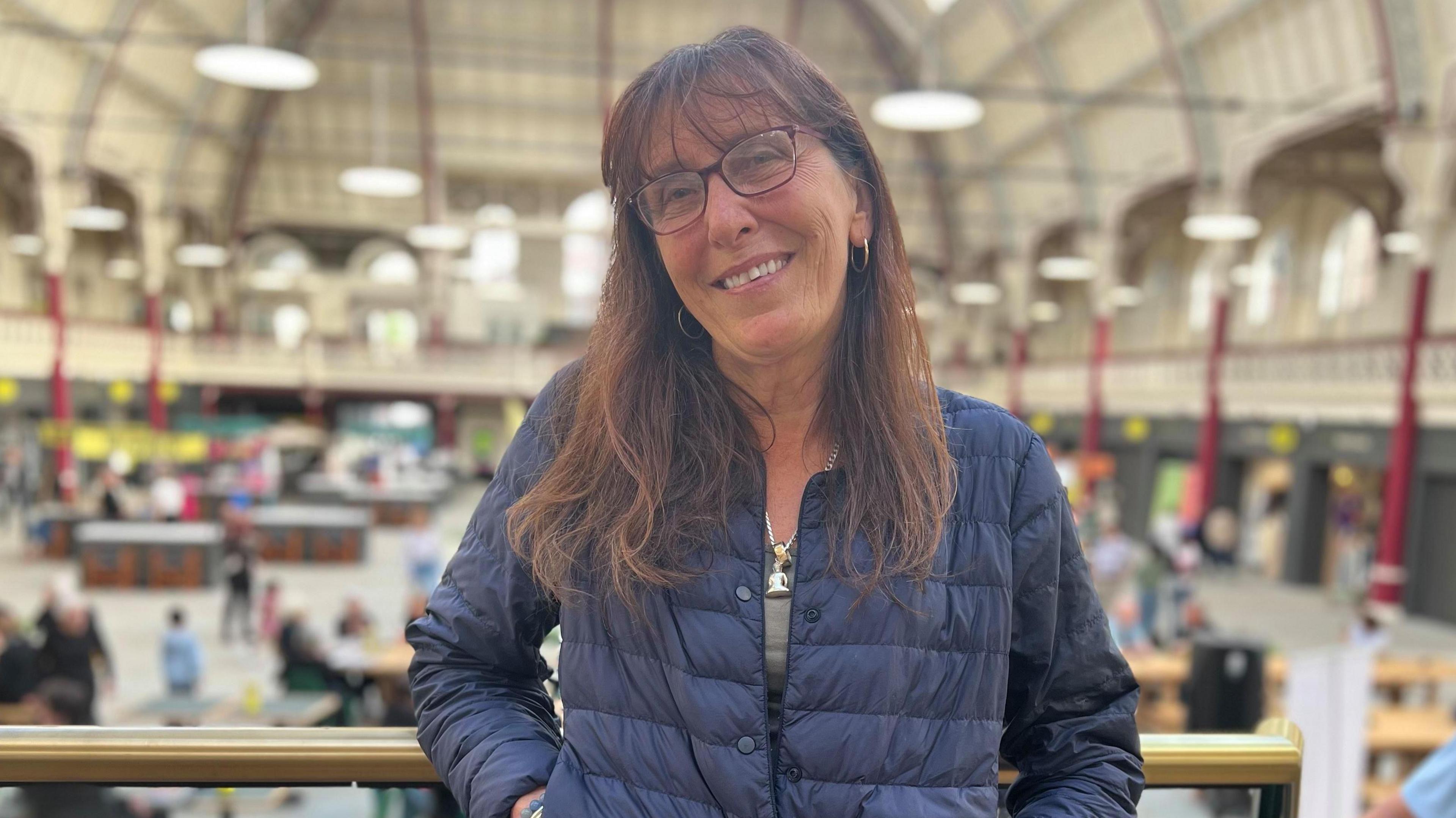Woman in blue puffer jacket, long brown hair and glasses standing on the balcony of a Victorian building with market stalls on the floor below