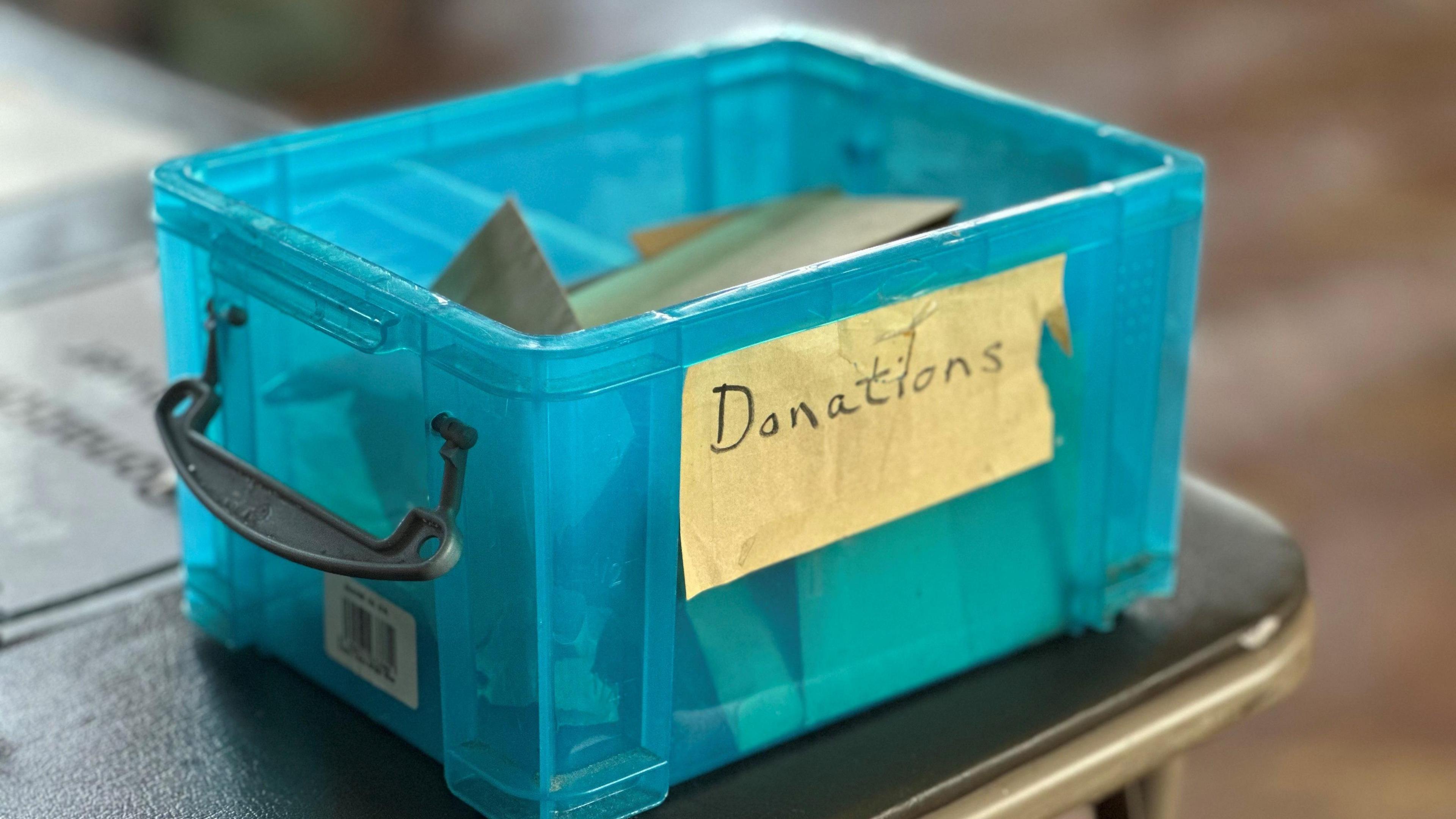 A blue plastic box with handles filled with small brown envelopes sitting on the edge of a table. It has a yellow note stuck to the side with the word 'Donations' written on it.