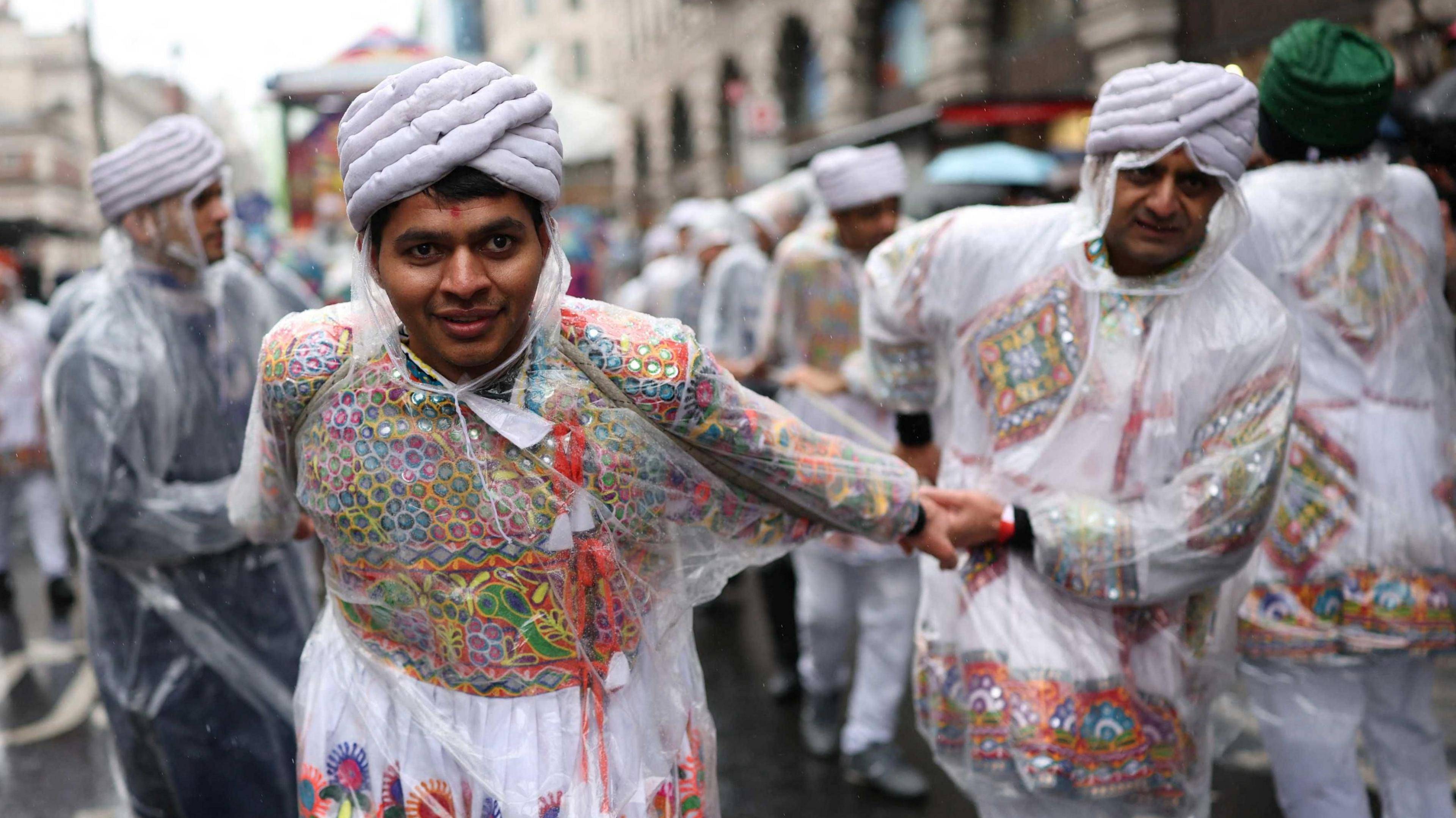 Two men in traditional Indian clothes, wearing rain ponchos, dance in the rain. 