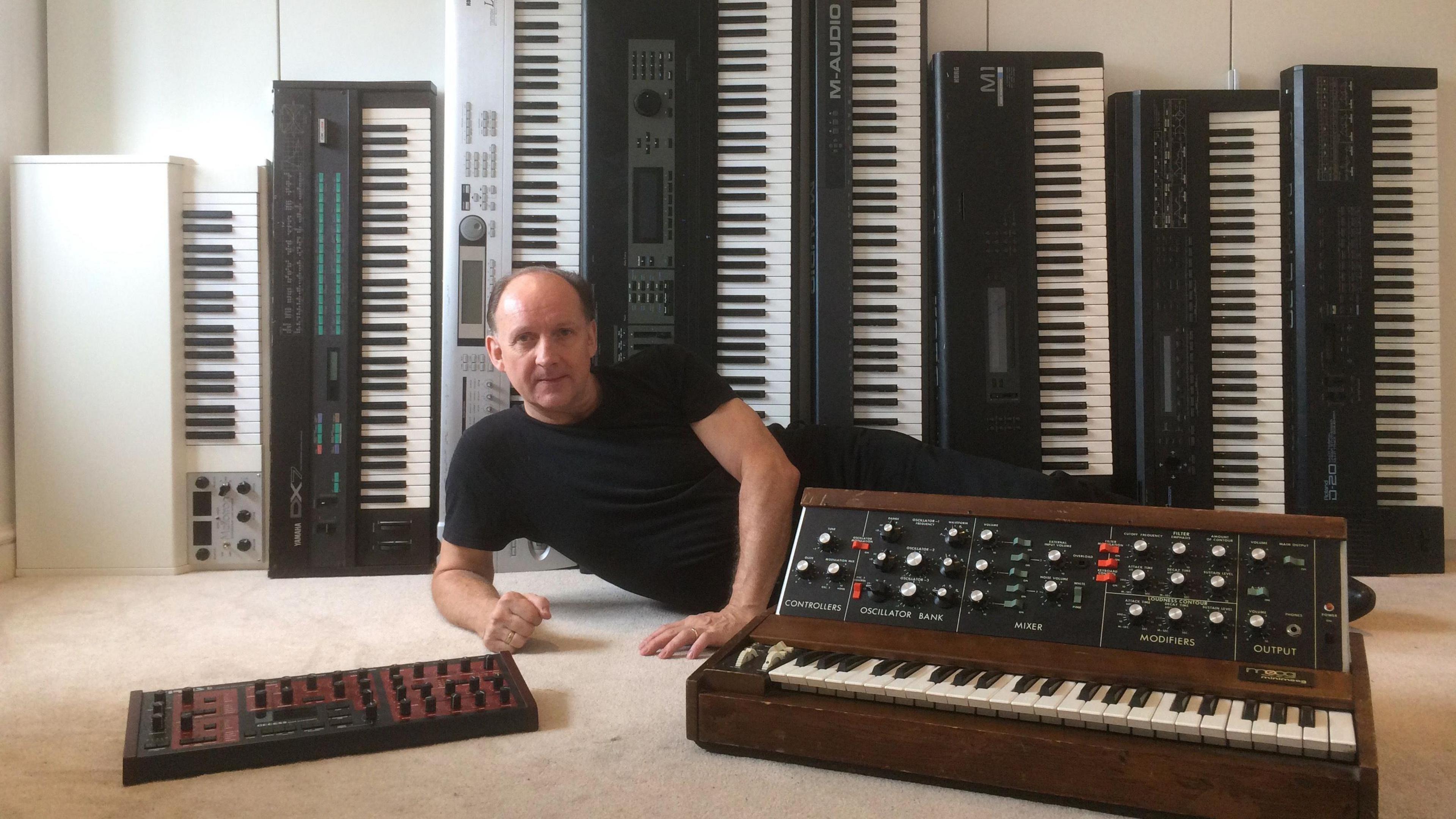 This picture was taken prior to Mike contracting necrotising fasciitis. He is lying on the floor, looking into the camera, leaning on his left arm, surrounded by 9 different keyboards and a studio mixer.