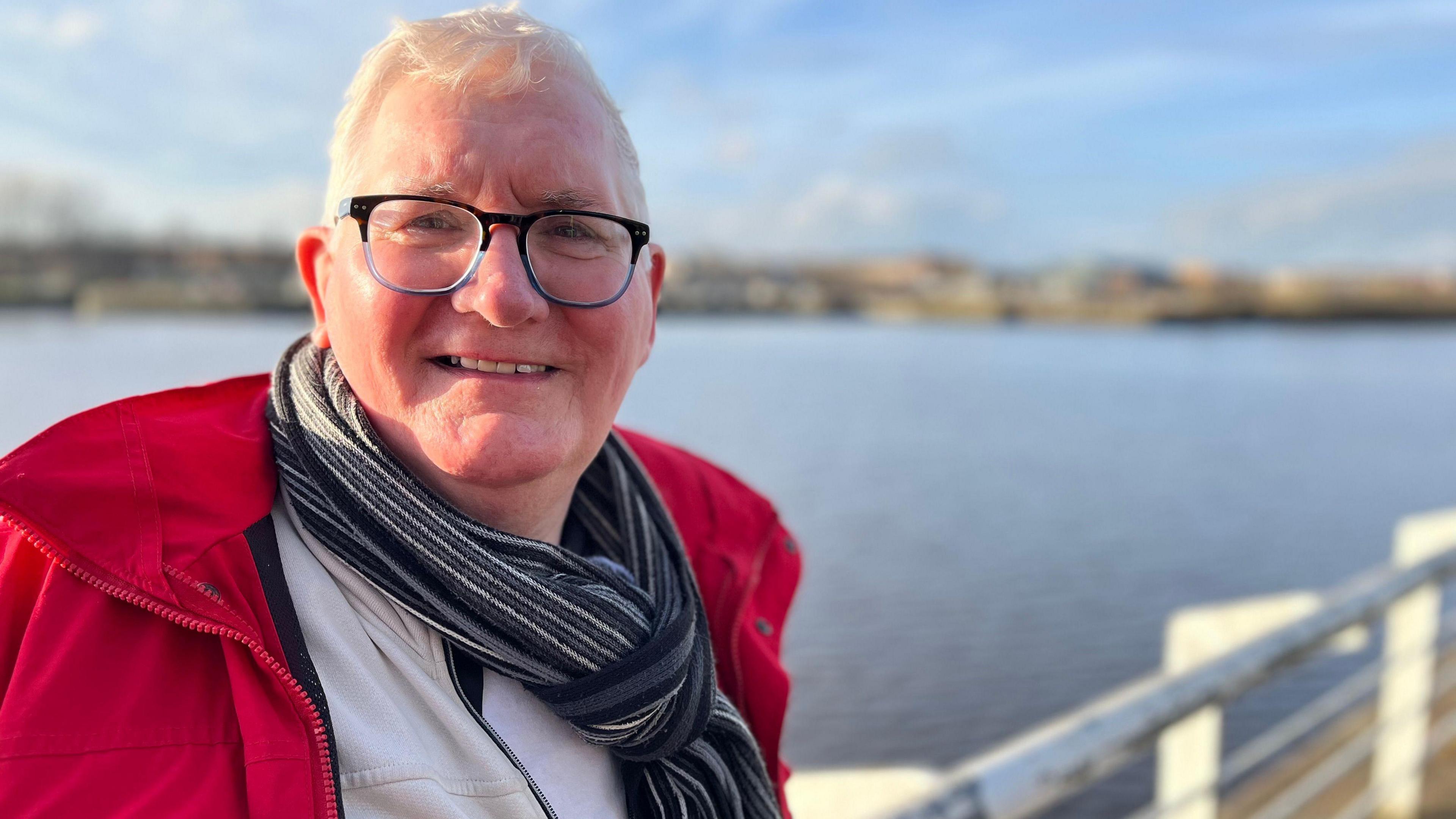 John Karol looking right at the camera. He is wearing a red jacket on top of a light-coloured top and a grey and black scarf tied round his neck. He has dark-rimmed glasses on and white hair. He is standing in front of the River Clyde, which is blurred in the background.