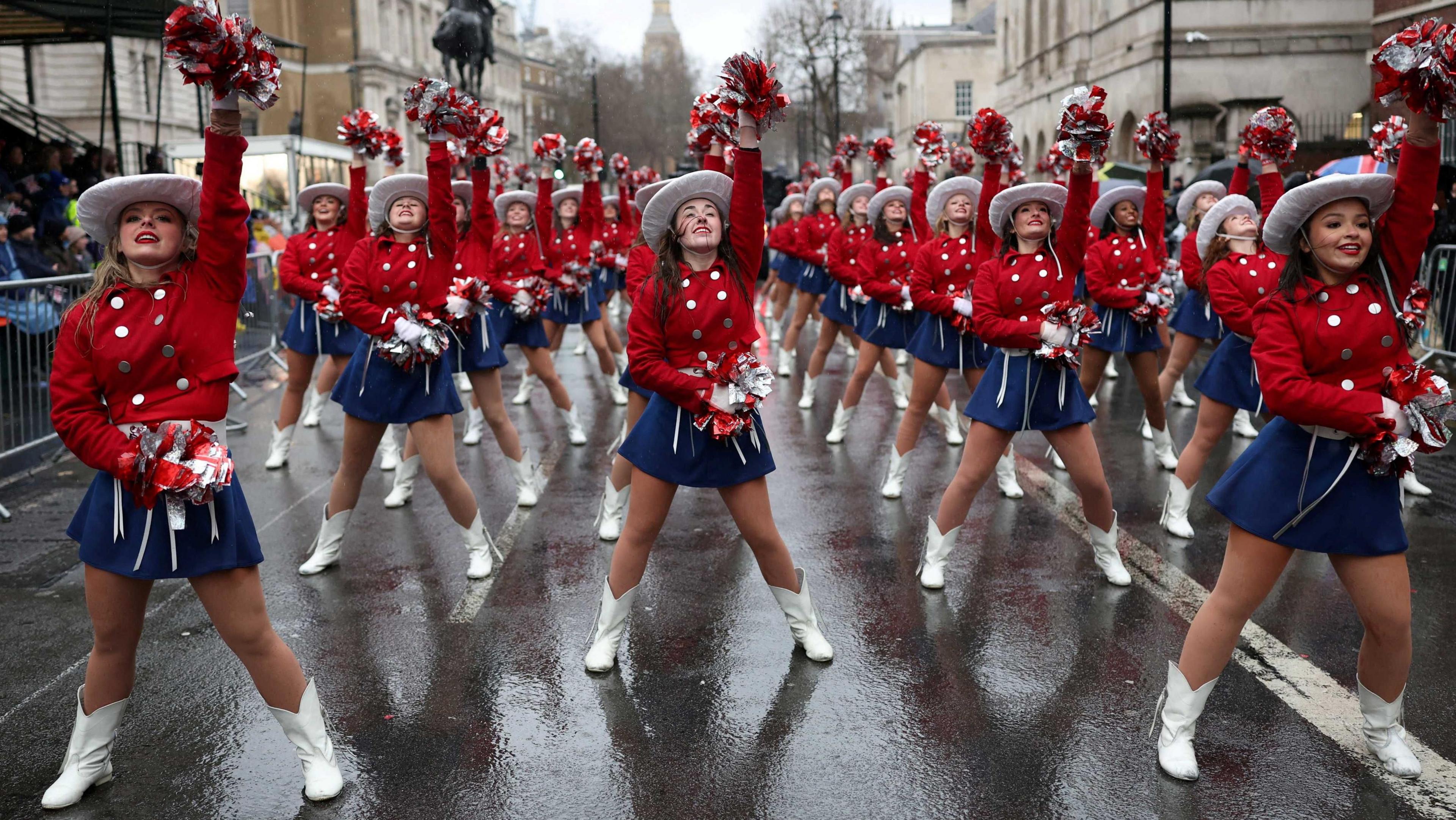 Three rows of female cheerleaders wearing white boots, blue skirts, red blouses with silver buttons and white Stetson hats, hold aloft their white pompoms. BIg Ben is visible in the background. 