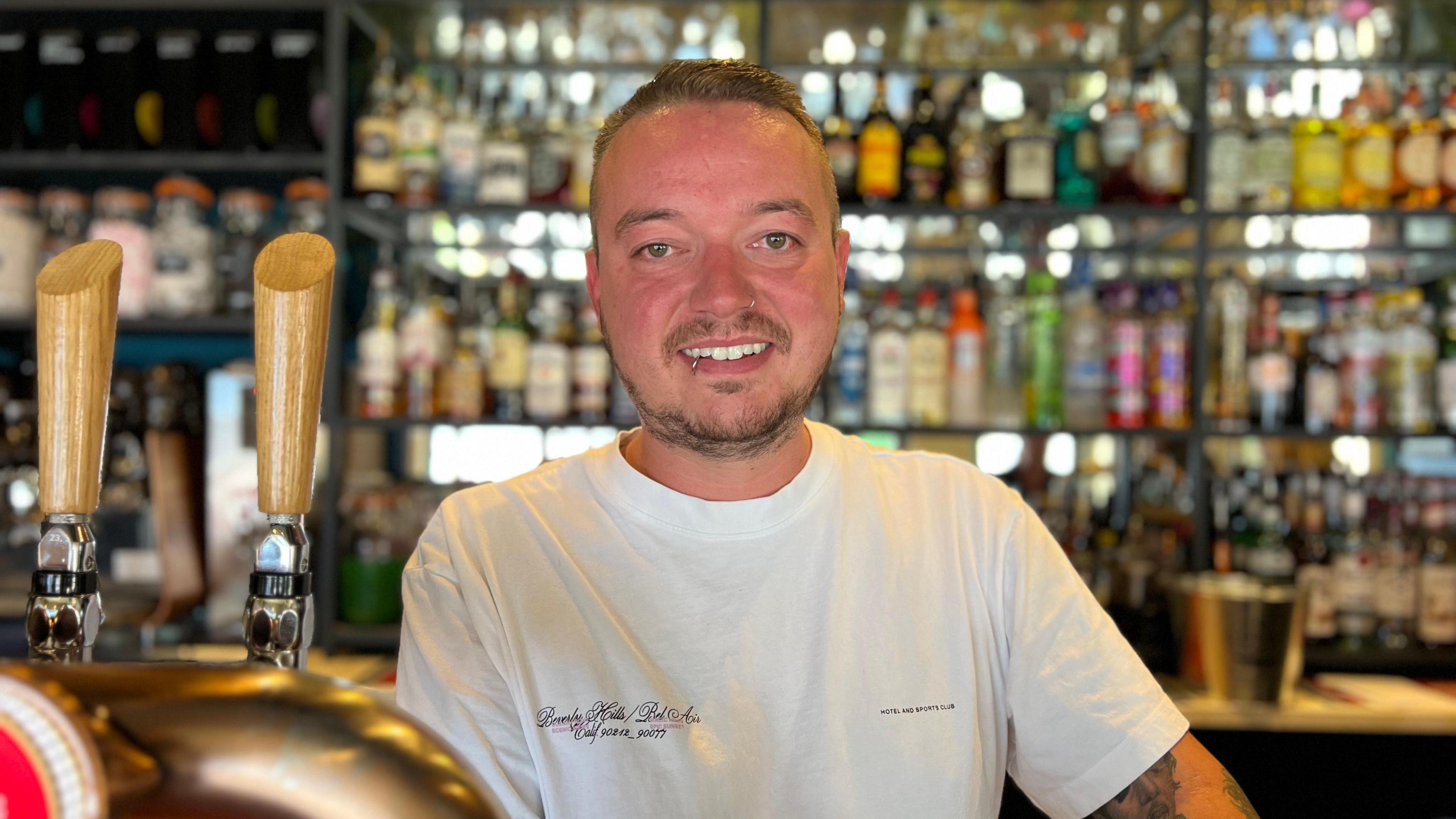 A man with short dark hair and a beard and a pierced nose and lip. He is wearing a white T-shirt and is standing behind a bar with beer pumps. There are bottles on a shelf in the background.