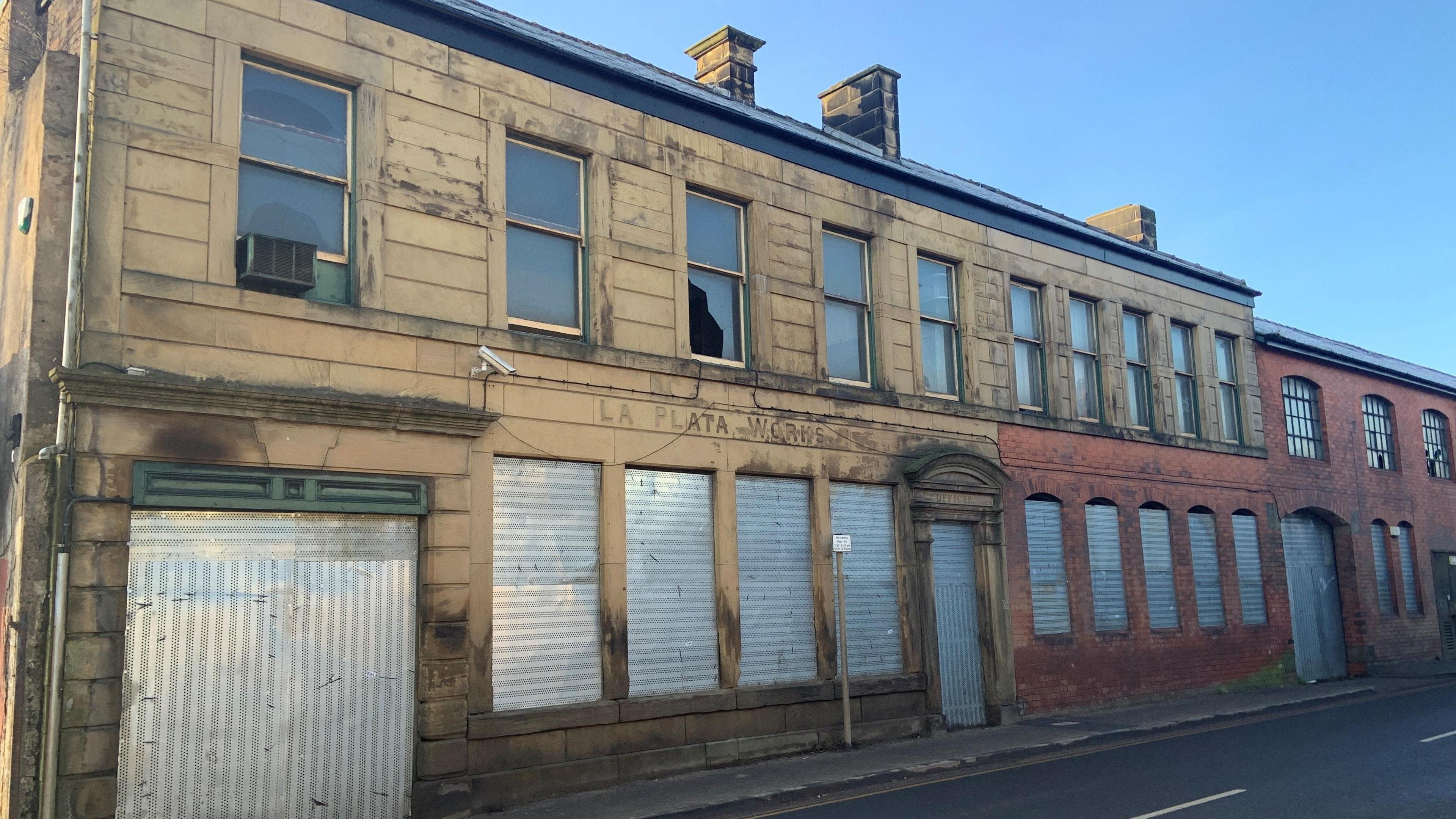 A former manufacturing building is a mix of brown stone and red brick. The windows and gateway on the ground floor have metal shutters covering them. Some of the windows on the first floor are broken. The words La Plata Works are carved into the stone work at the front of the building