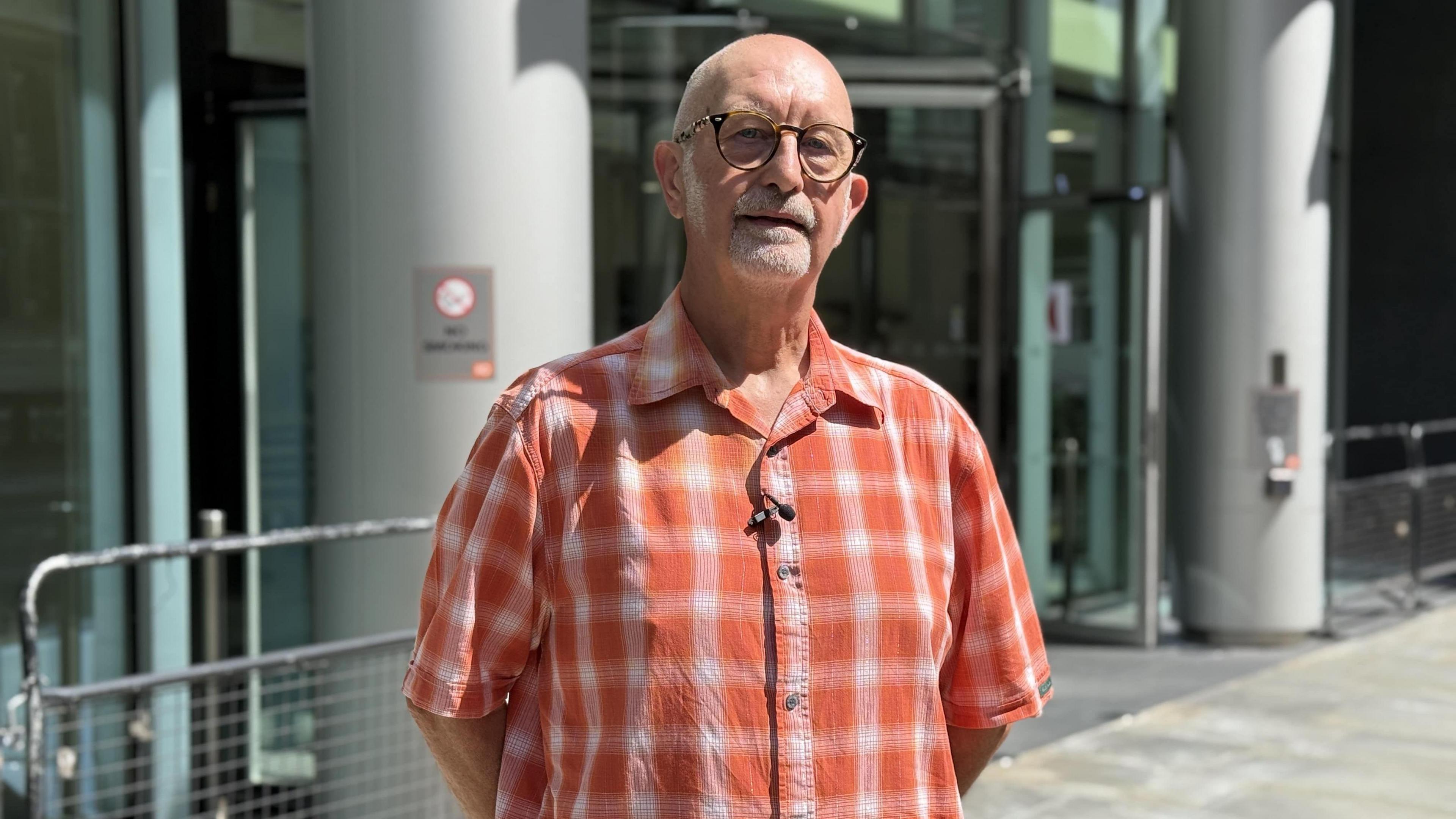 Graham Lewis has a short white beard and spectacles in an orange checked shirt. He is stood outside the modern-looking Rolls Building in London on a sunny day.