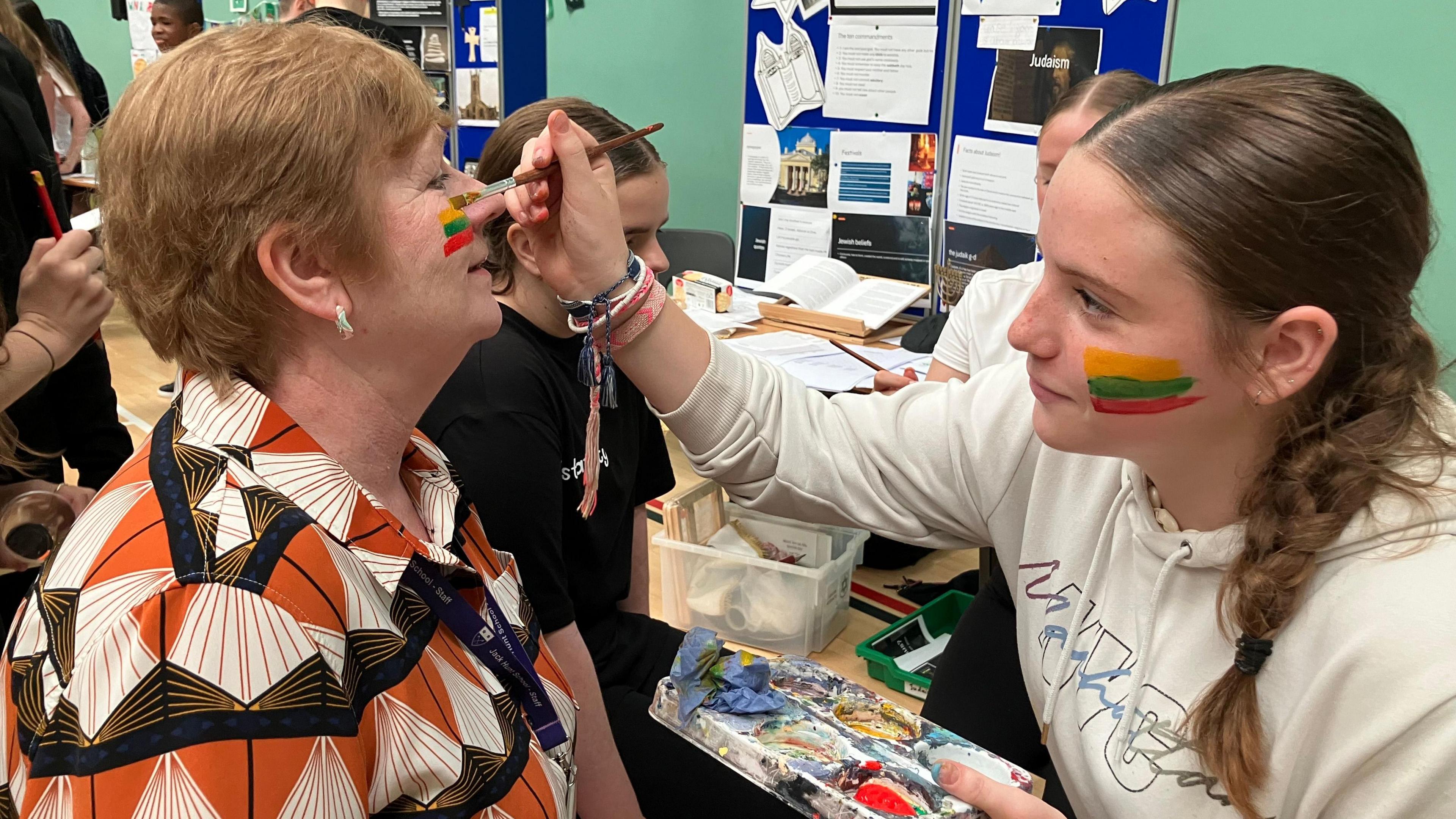 A girl painting a woman's face with Lithuanian flag colours - yellow, green and red 