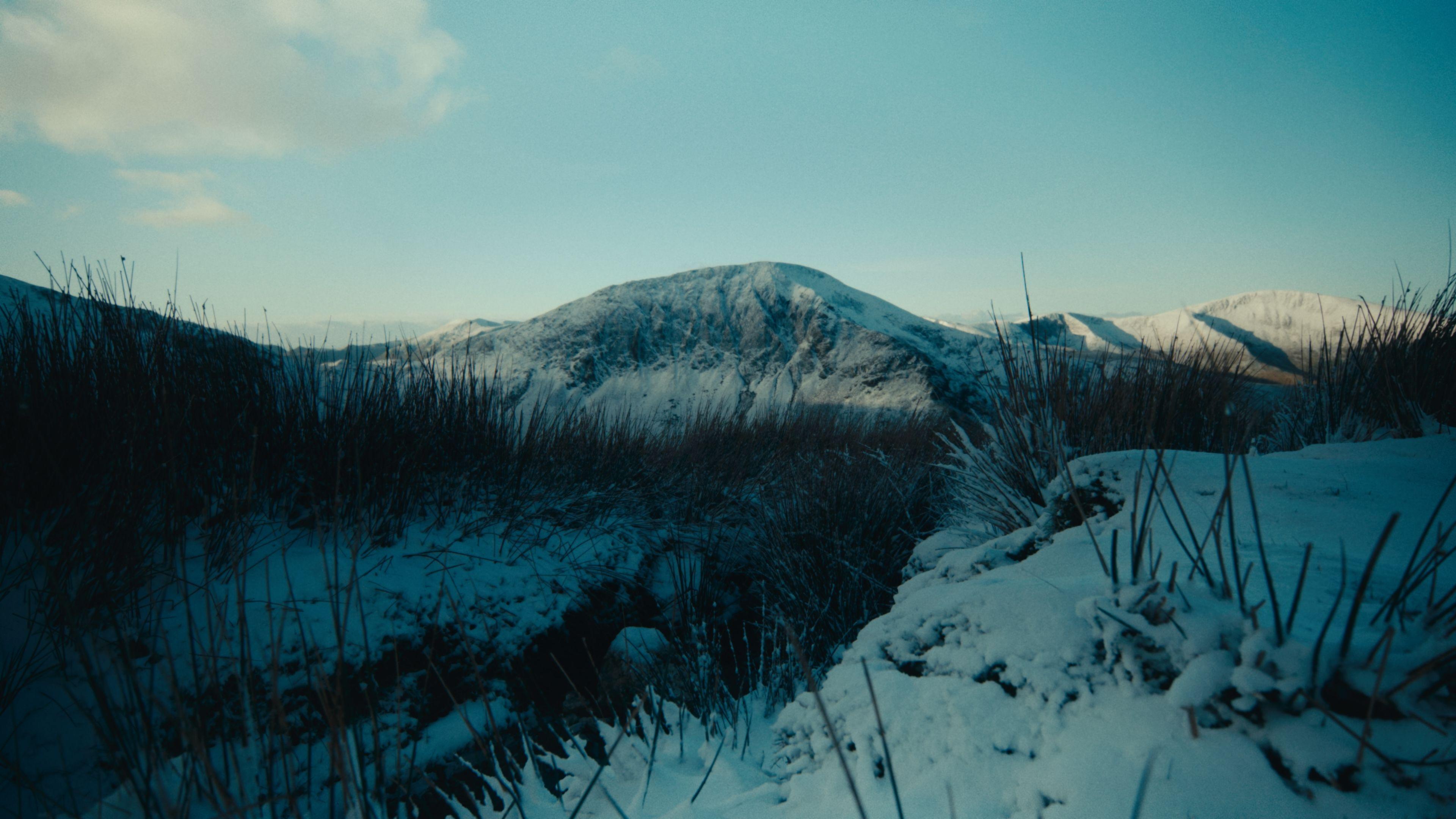 An icy cold snowdonia. There is a snow-covered mountain in the distance and frosty grass in the foreground. It's sunny.