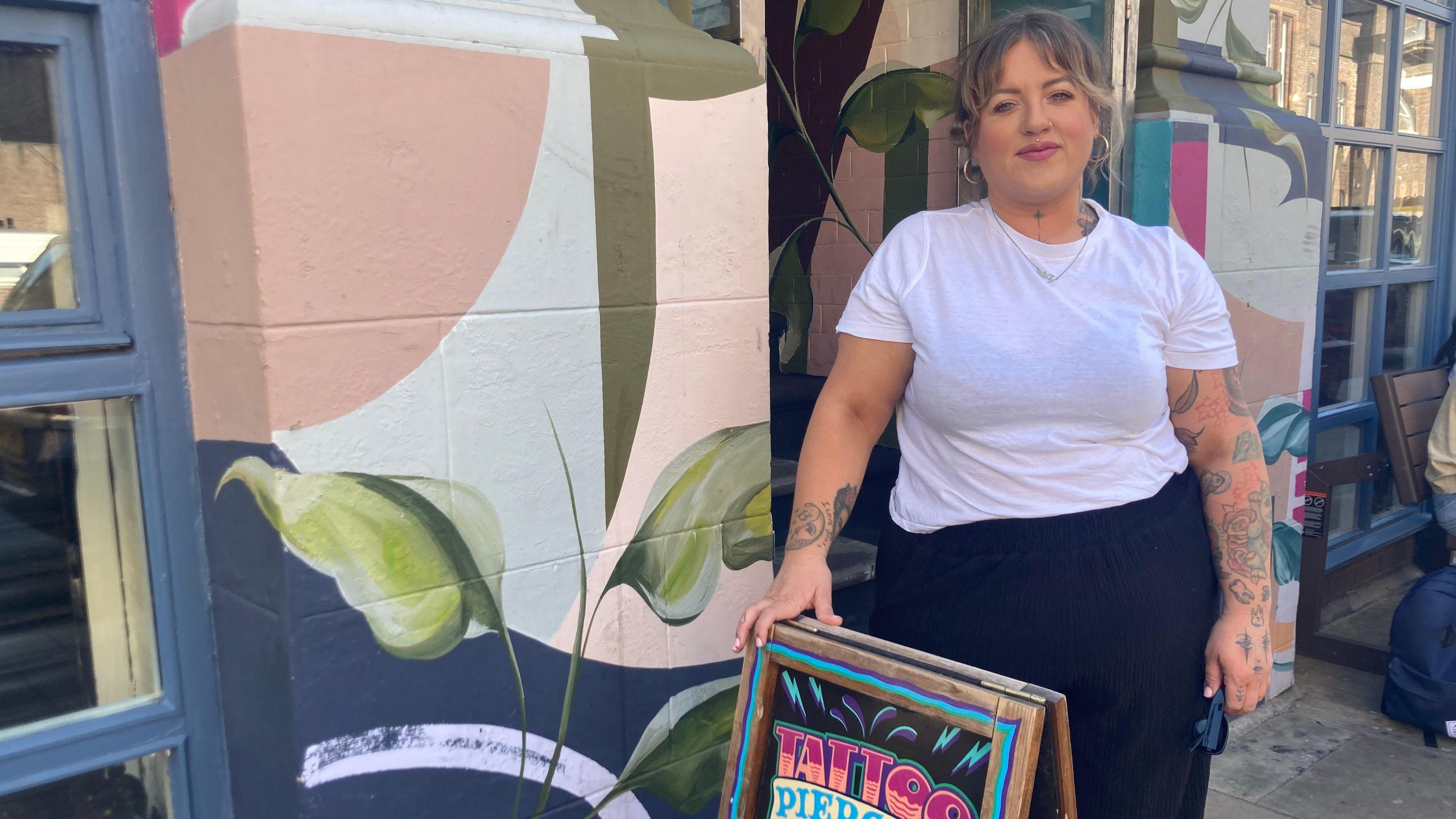 Hollie stands outside the tattoo parlour, it has a flora mural painted on the entrance. She wears a white t-shirt and black trousers. She has a sleeve of tattoos on her left arm.