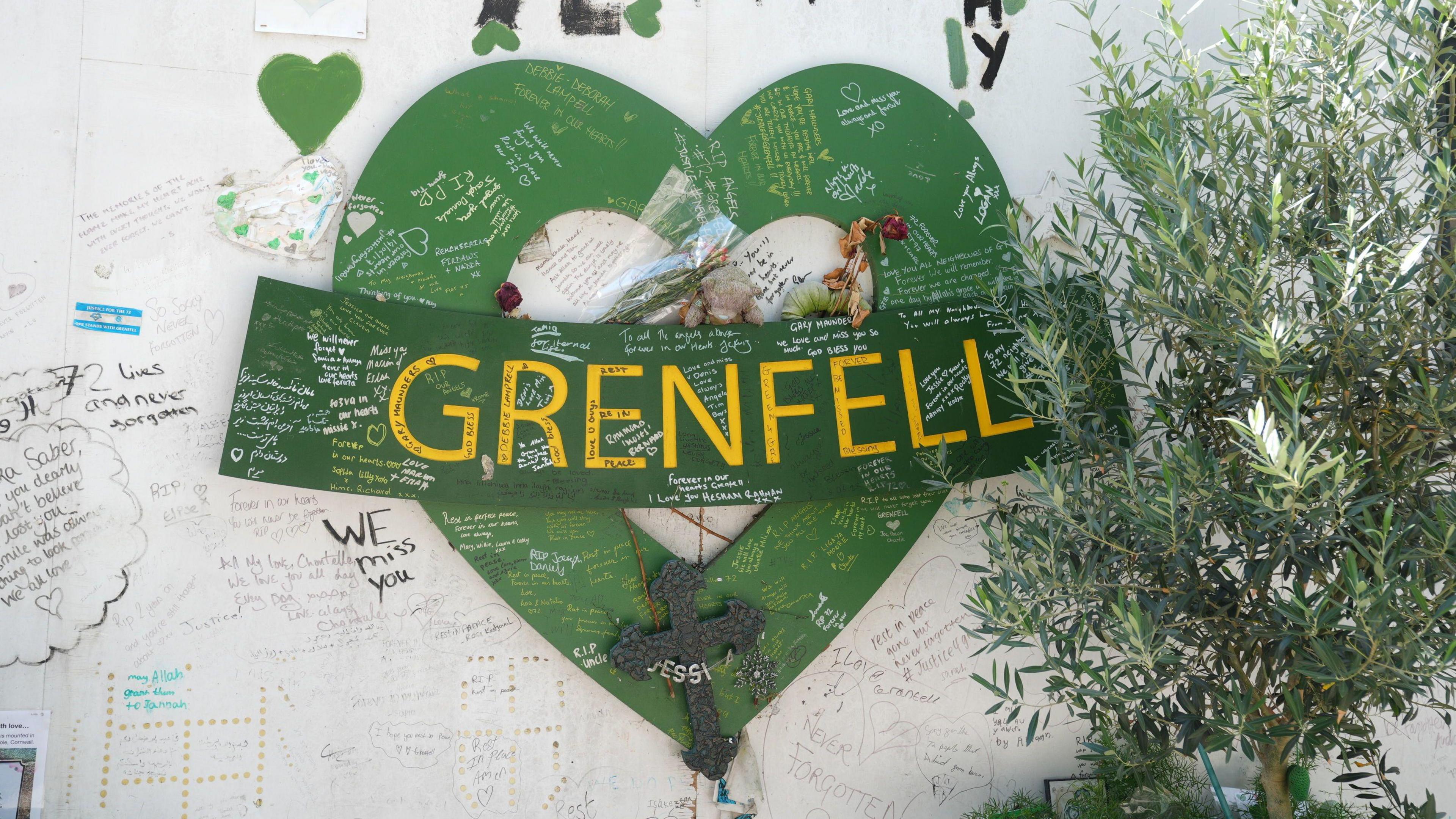 A view of the grenfell memorial wall, with a large green heart with a banner across it saying 'Grenfell' in yellow letters. There are handwritten messages all over the heart and the white wall behind it. An olive tree is planted to the right of the heart