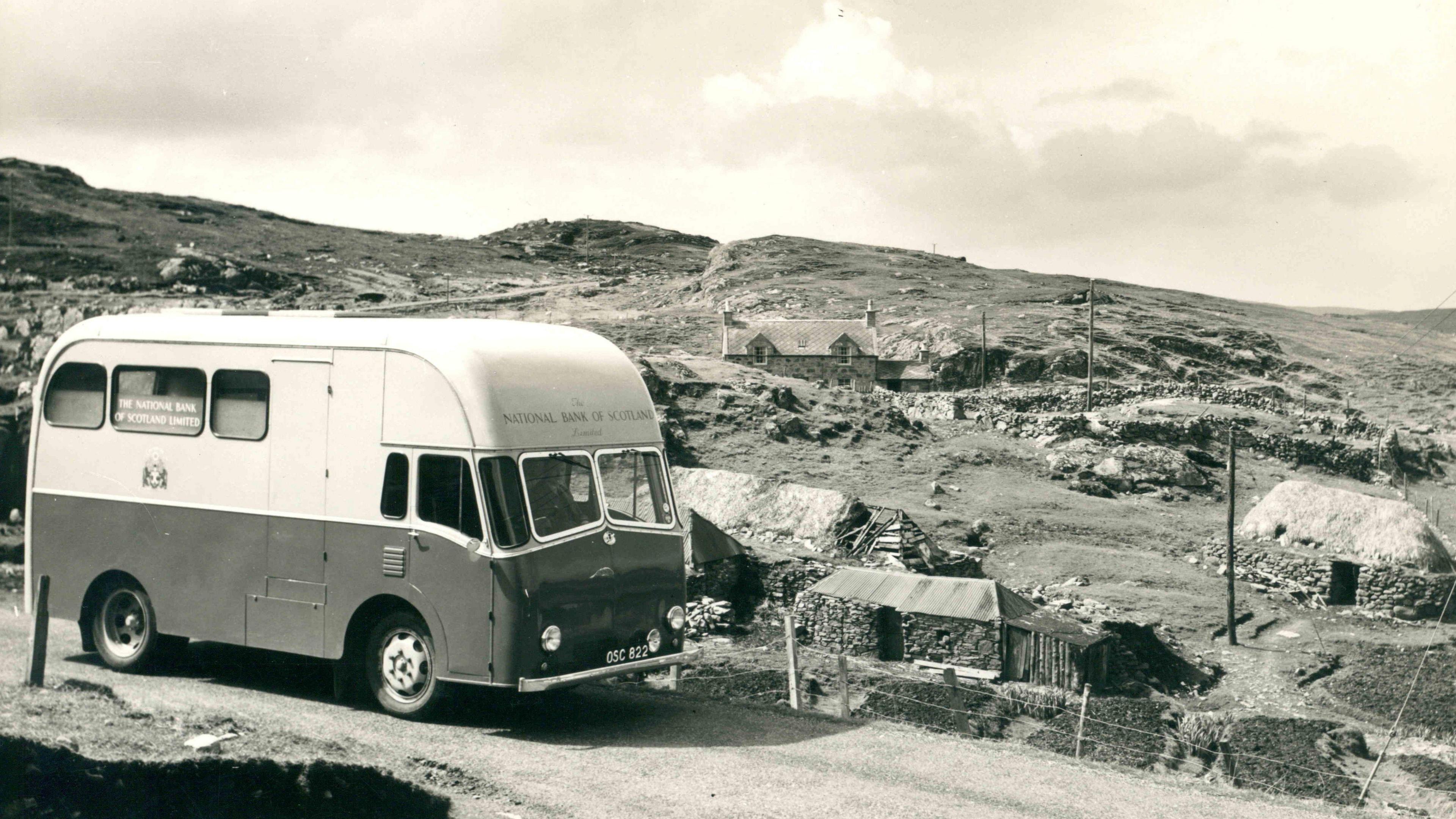 Dealbh dubh is geal de bhana Banca Rìoghail na h-Alba ann am baile beag Gàidhealach. Tha taighean-tughaidh ann cho math ri taigh-croite nas ùire. 