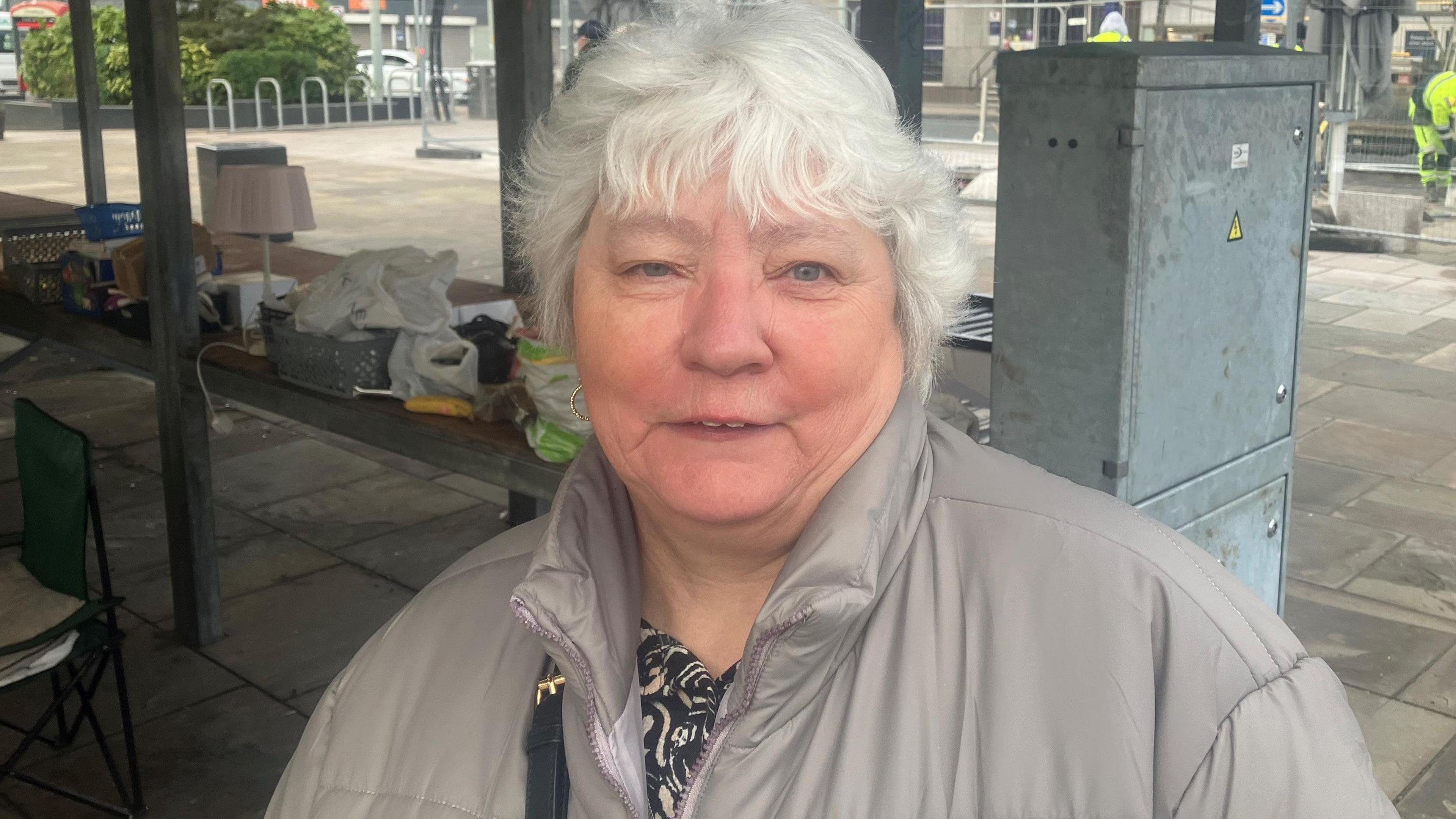 Louise Aliceto with short white hair wearing a hoop earrings and a black and white blouse with a grey coat in Hyde town centre