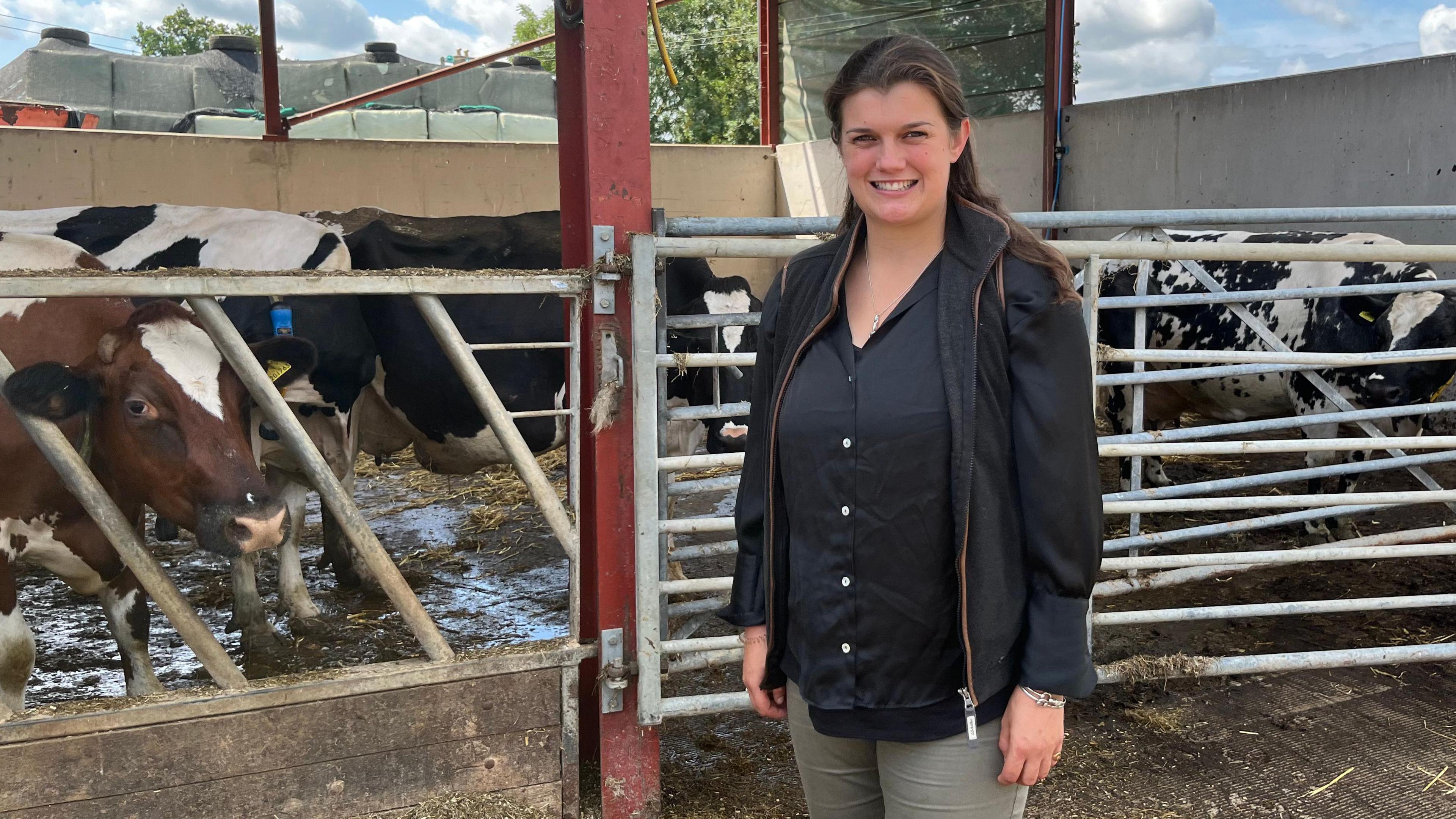 A young woman with long brown hair smiles as she stands in front of a shed full of cows. She is wearing a black shirt, a black waistcoat, and grey trousers.