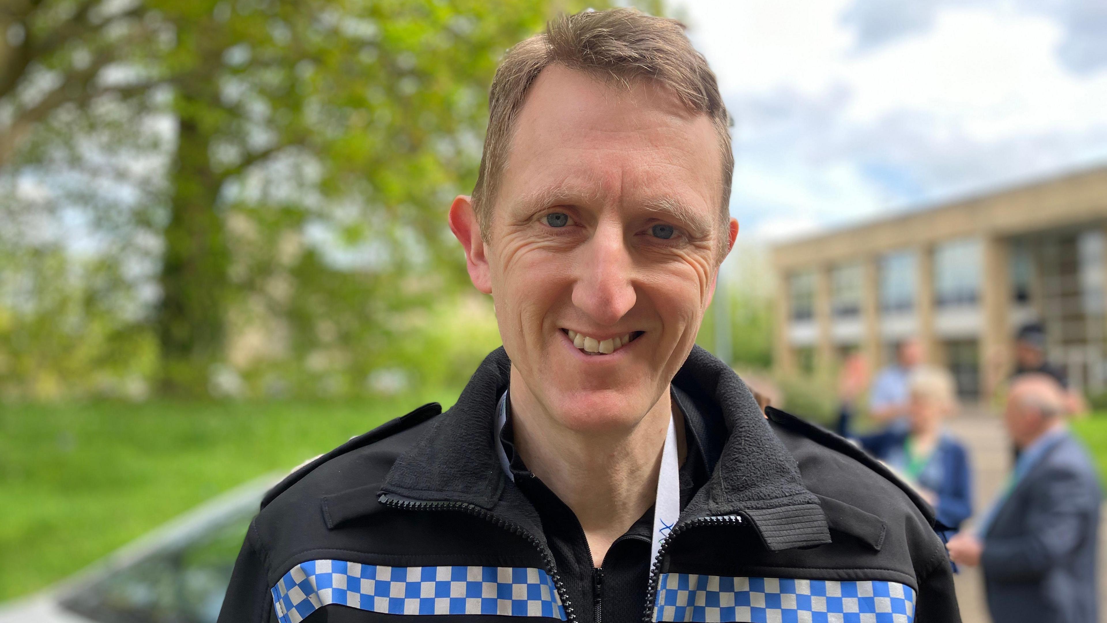 Mr Cooper has light brown hair and is smiling at the camera. He is wearing black police uniform with a silver and blue reflective strip across the chest