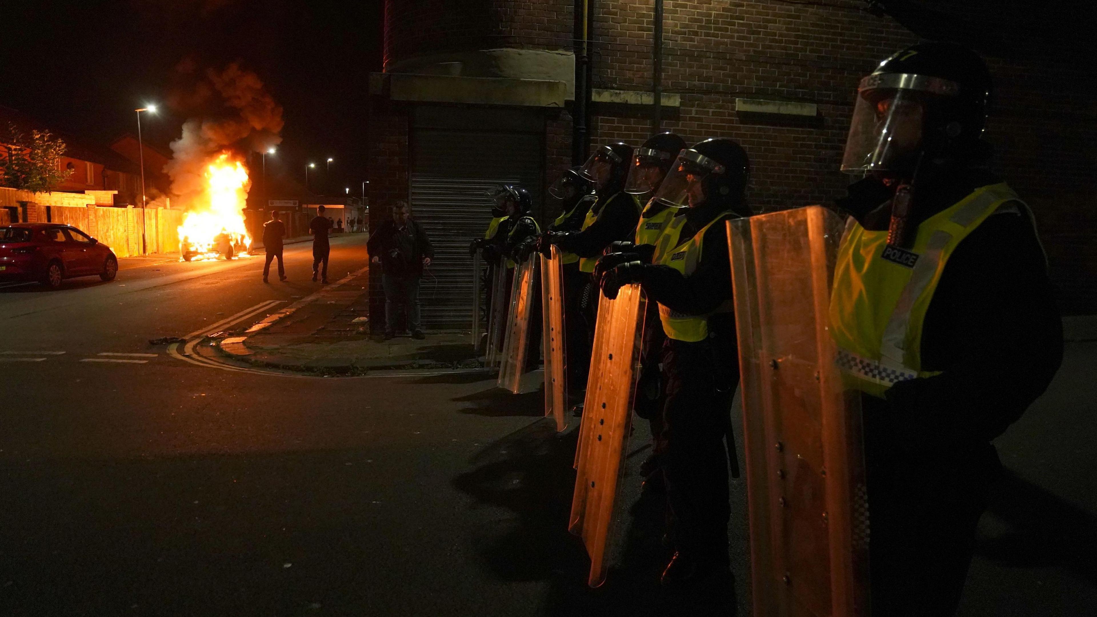 Police officers lined up in the dark, wearing riot gear and holding shields, with a burning car in the distance.