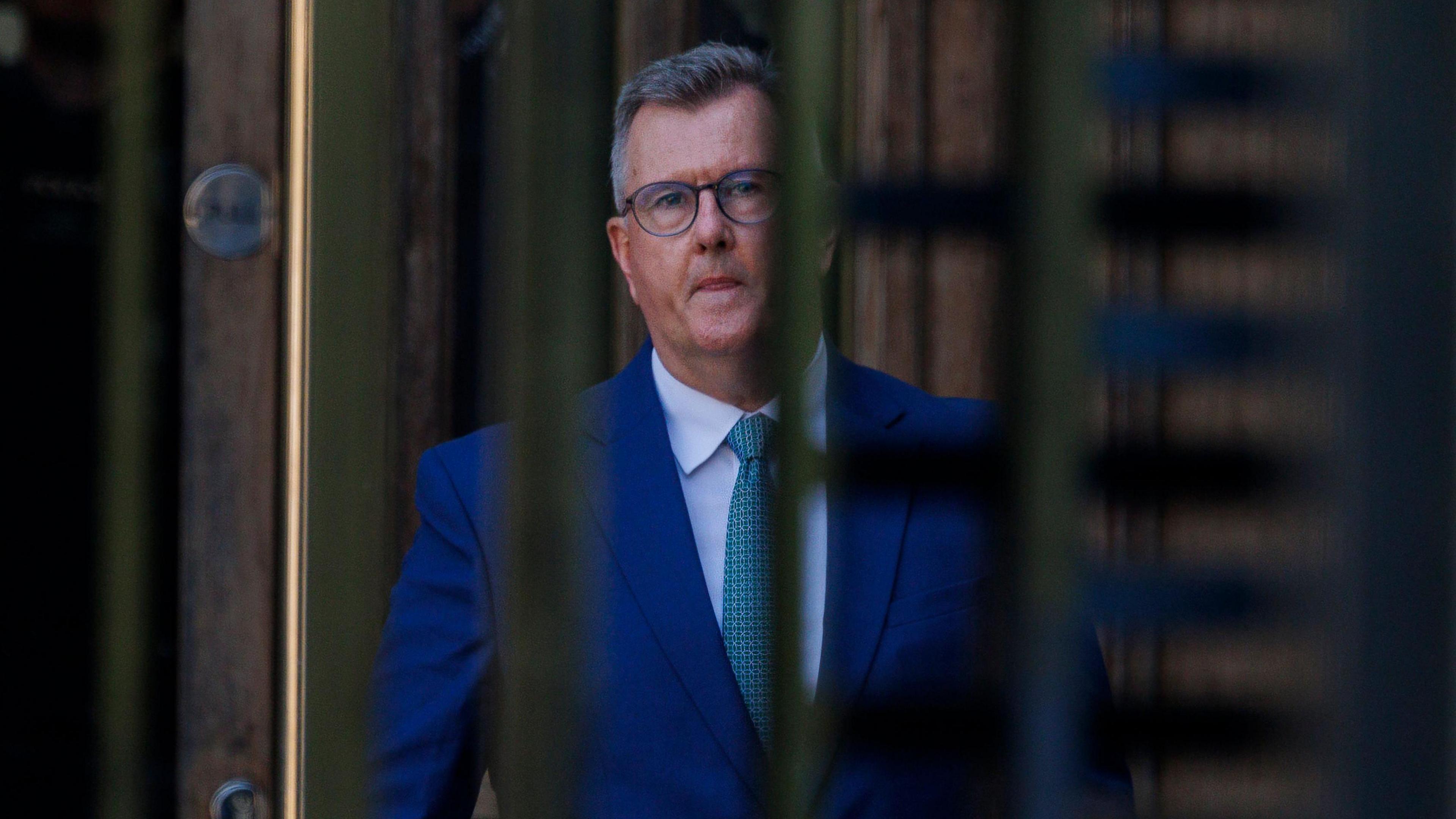 Jeffrey Donaldson, a man in a blue suit with short grey hair and glasses, seen through the bars of an entrance gate.