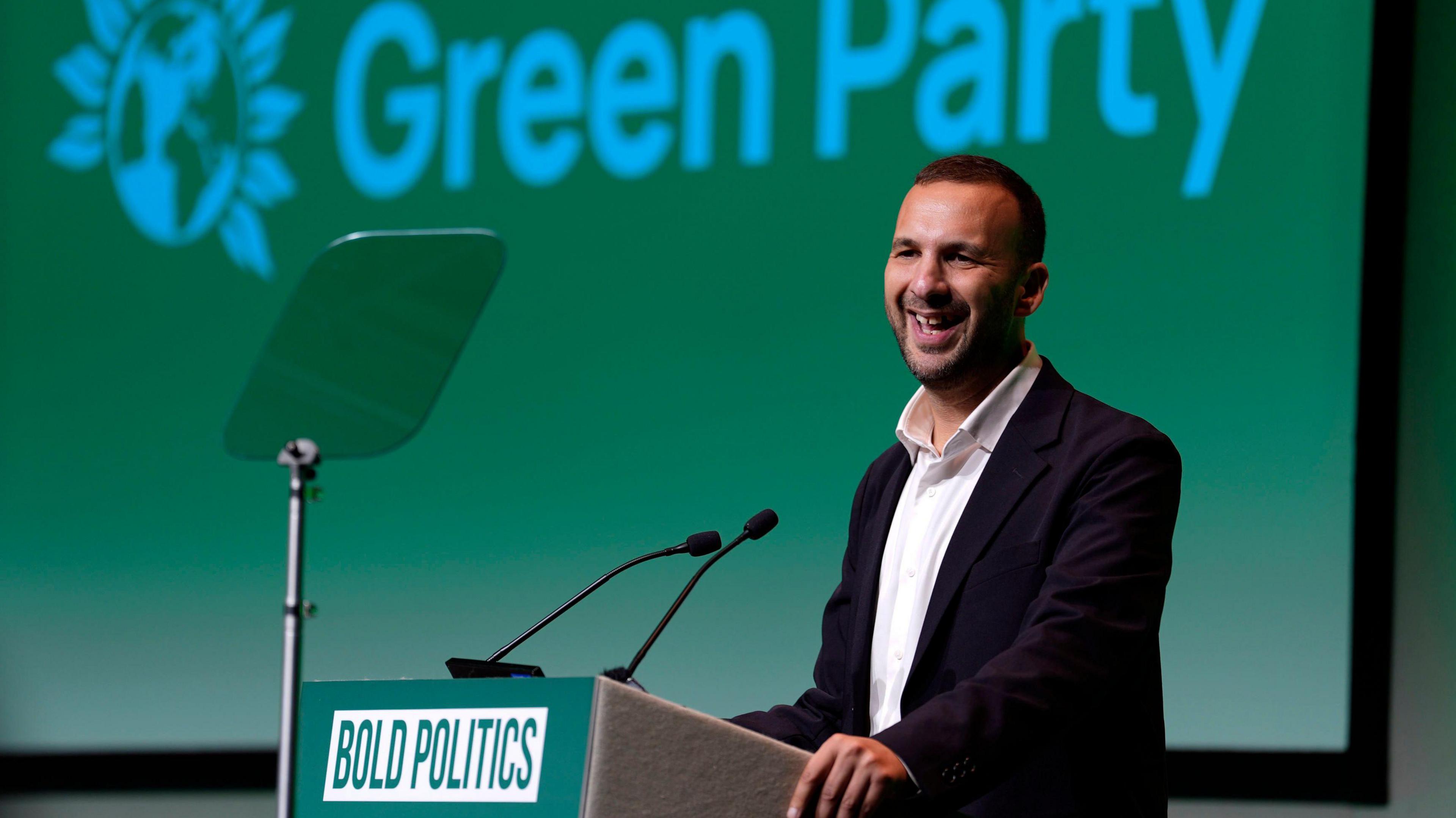 Zack Polanski in a white shirt and black jacket standing at a podium with 'Green Party' on a screen behind him and the word 'bold politics' on the podium.