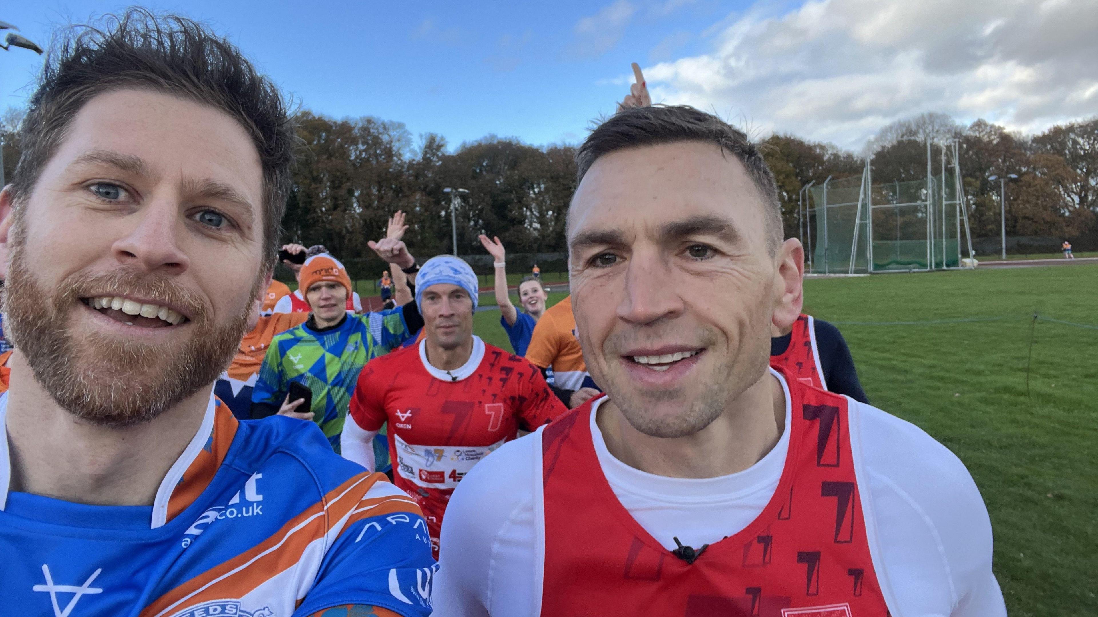 Sam has brown hair and is wearing a blue, orange and white motor neurone disease foundation t-shirt. He is taking a selfie with Kevin Sinfield, who has short brown hair and is wearing a white top with a red MND foundation vest on top. In the background are other runners waving to the camera.