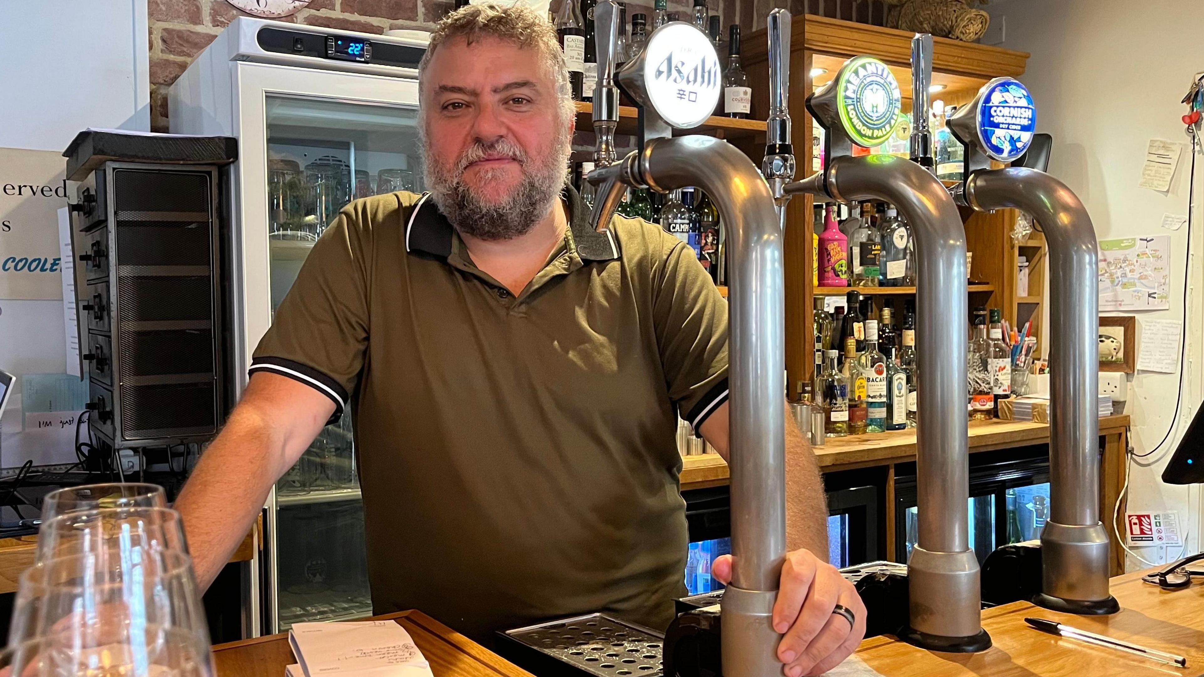 Daniel Bond, wearing a khaki polo shirt, poses behind the bar at his restaurant. He stands behind three beer pumps, next to the wooden bar top.