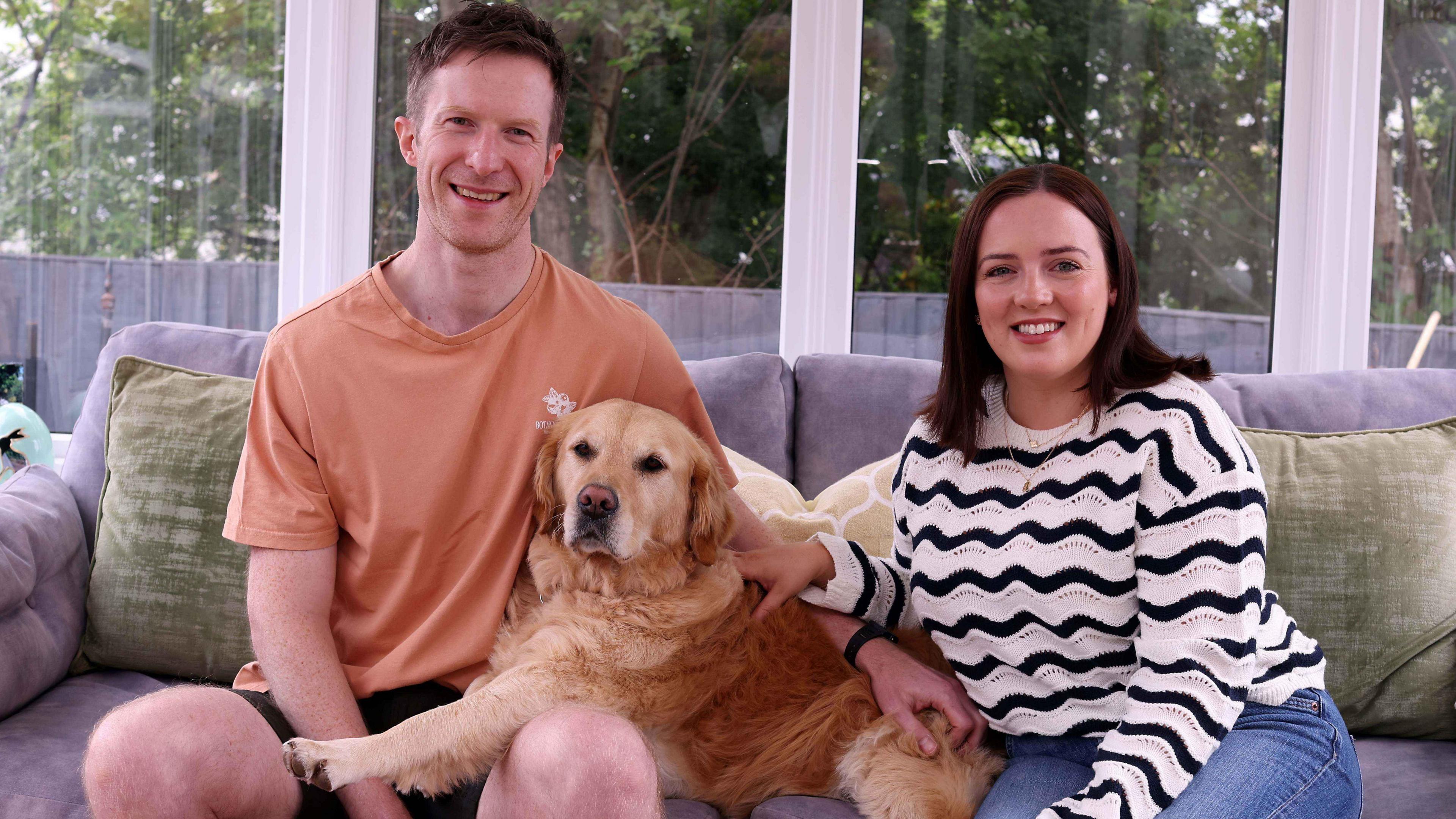 A man and woman sit on a grey couch, with a golden retriever dog between them. The man wears an orange t-shirt and black short. The woman wears blue jeans and a black and white striped jumper. Behind them are clear windows with white frames. Outside are green trees and a black fence, separating the trees from their garden 