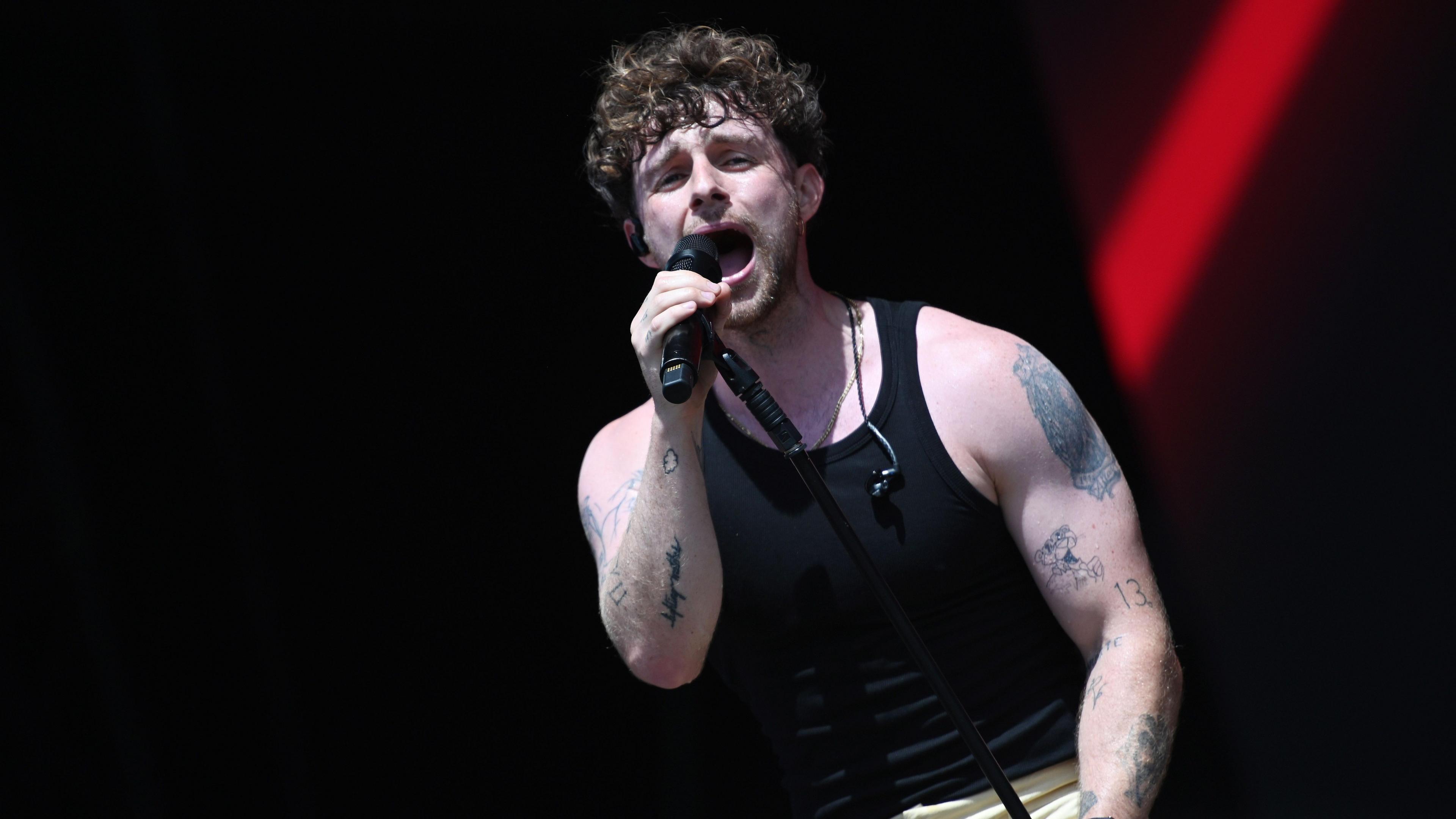Tom Grennan performing on stage in a black tank top and cream trousers.