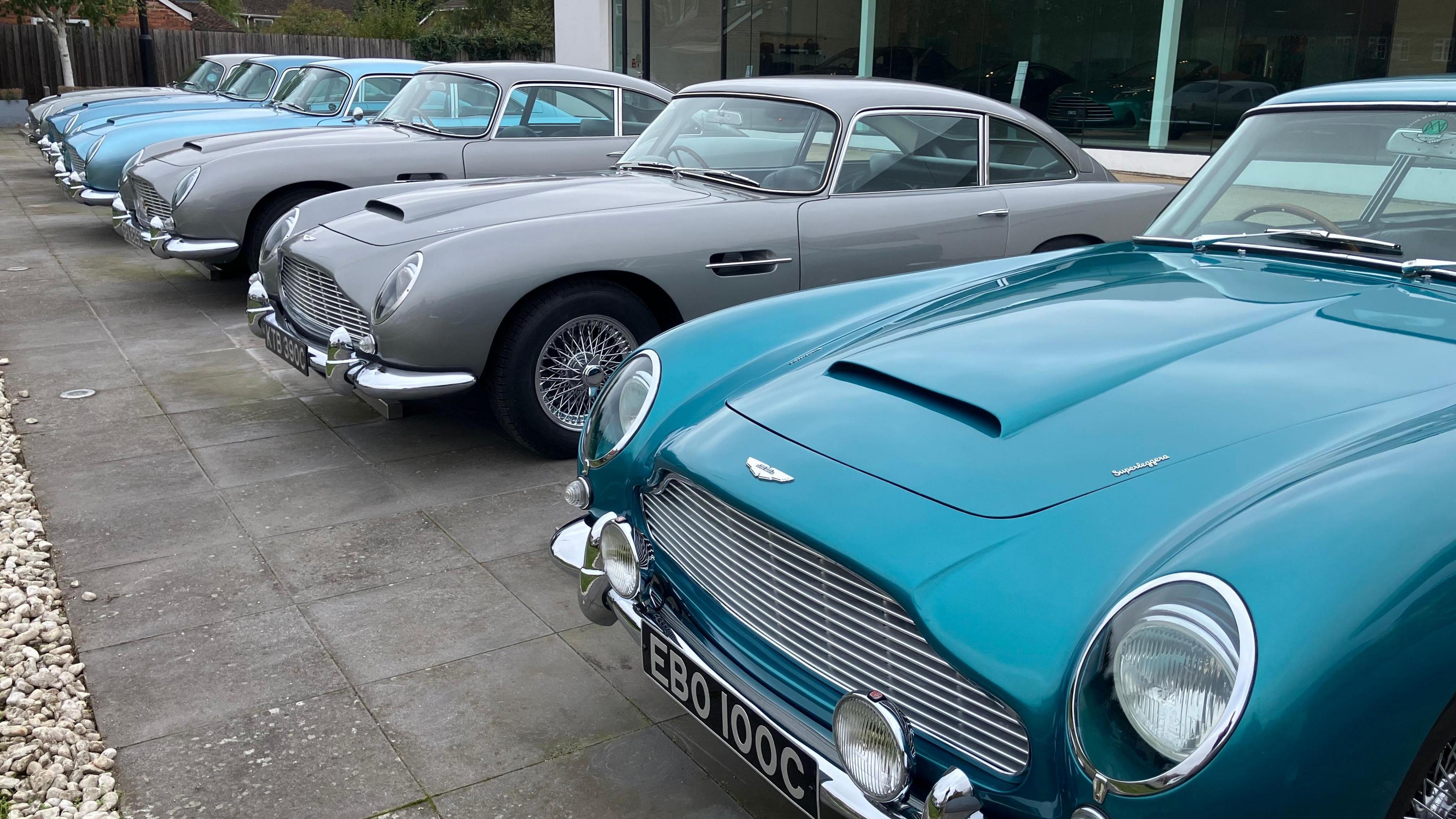 Six Aston Martin DB5s can be seen parked in a line in front of a glass fronted building. The nearest car to to the camera is a green-blue colour. They are parked on grey paving slabs.