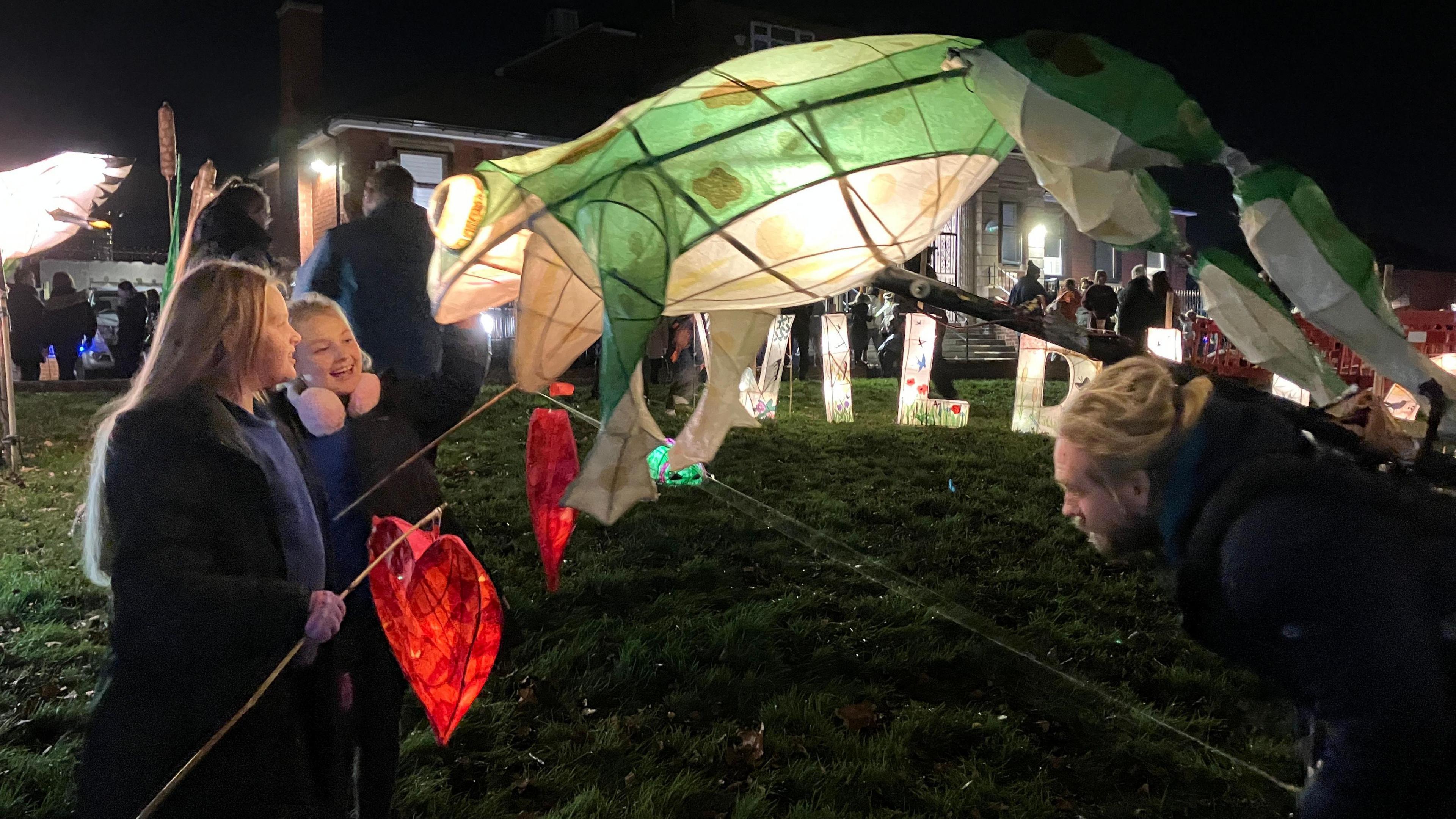 Two young girls look on as a puppeteer performs with a green light-up frog lantern.