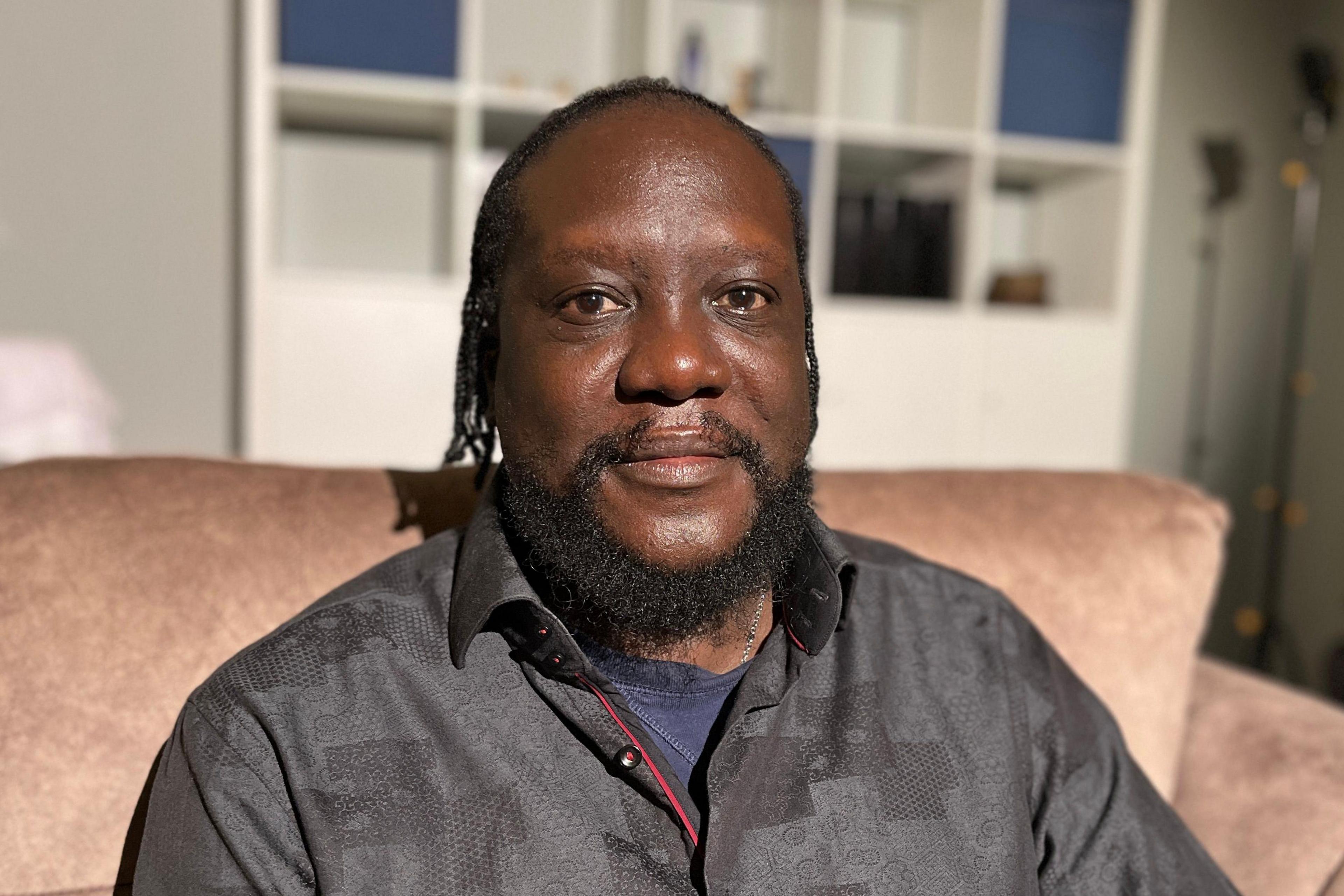 Henry Sabati McRae sitting on a pink sofa in his living room. Henry has brown eyes, longish hair and a beard. He wears a blue tee shirt and a black, textured, shirt