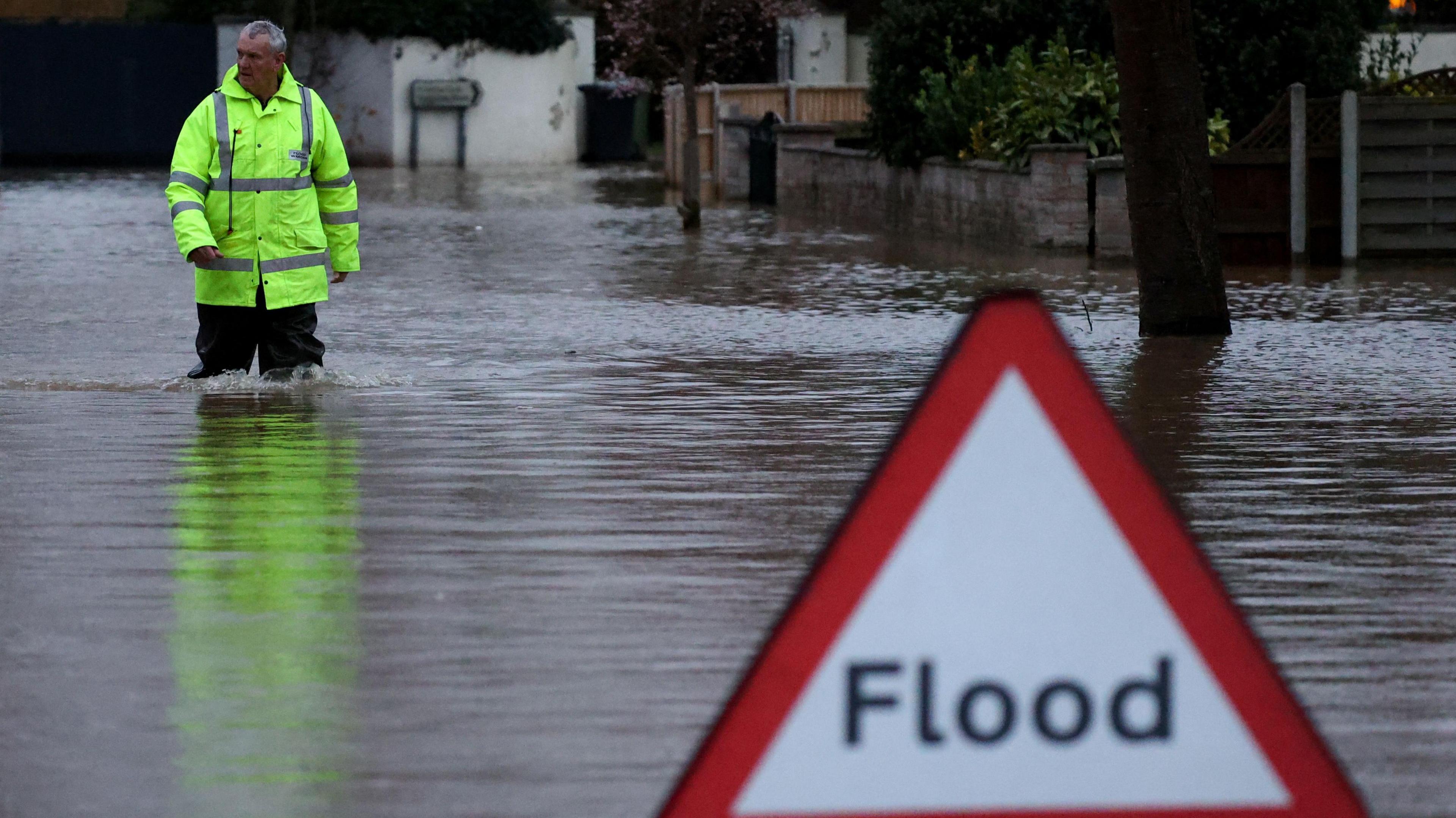 A triangular red sign that reads "flood" is pictured surrounded by water with a man in a high viz jacket walking behind. 