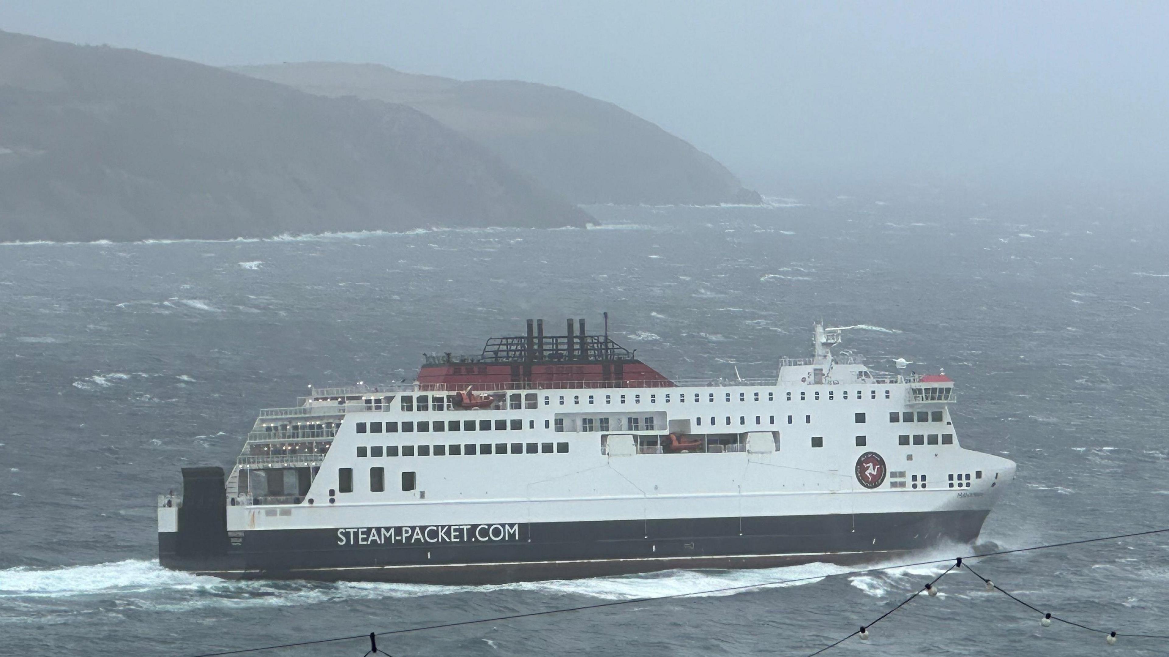 The Manxman ferry, which is red, white and black and has Steam-Packet.com on the side, sailing in Douglas Bay with a headland behind it. The sea around it is choppy.