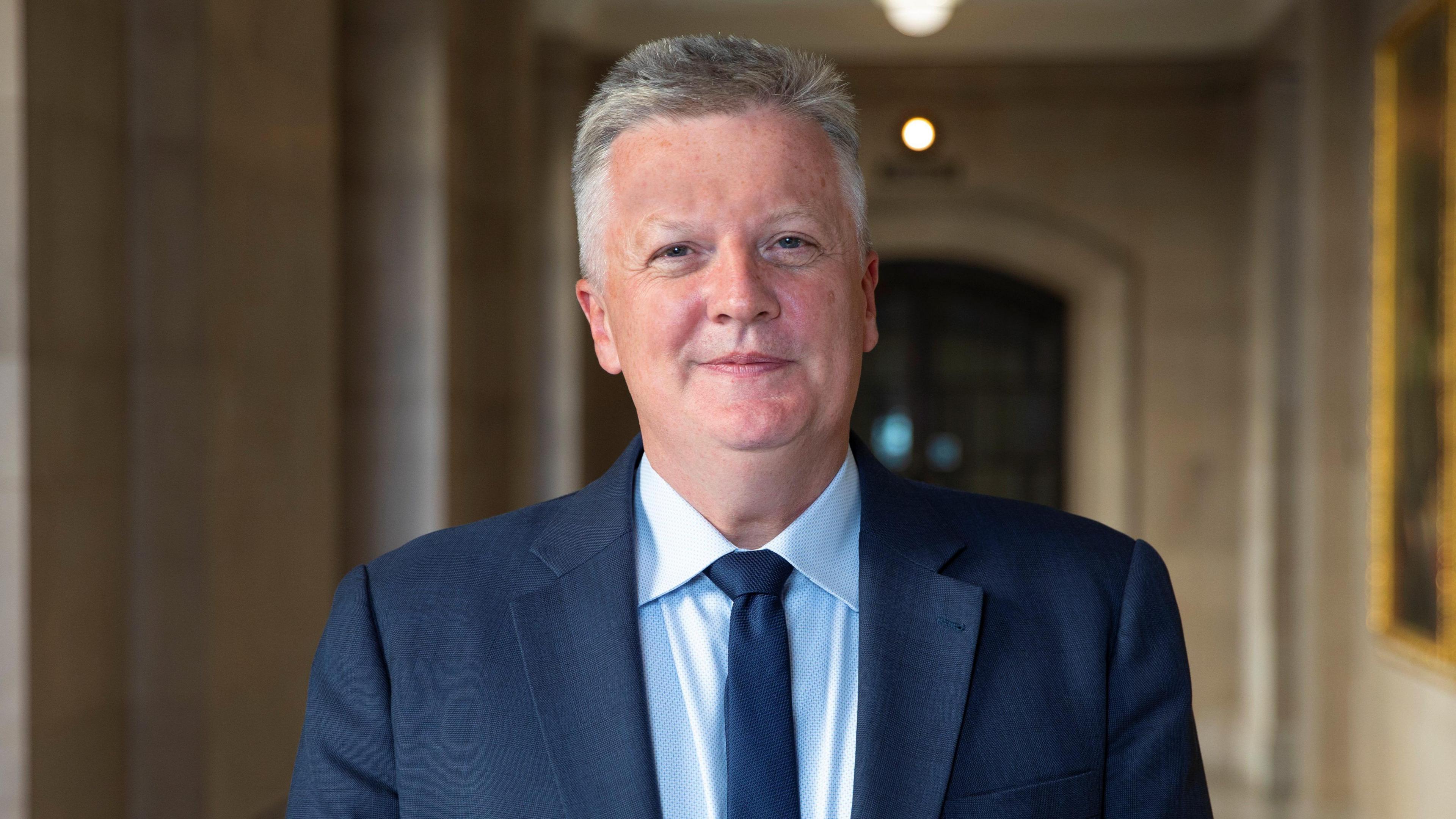 A man with short grey hair in a navy suit with a blue shirt and tie