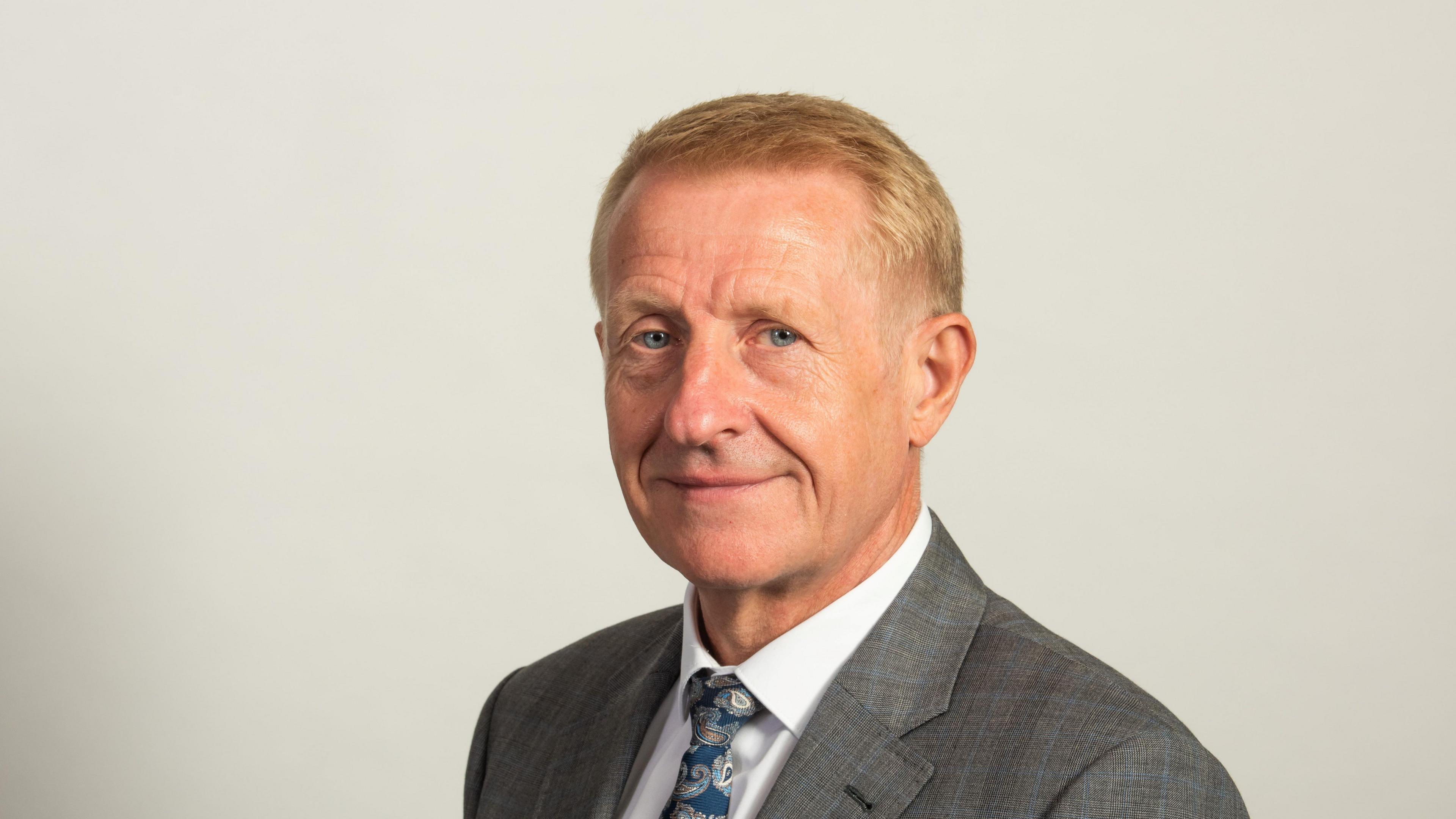 A man with short blonde hair wearing a grey suit, white shirt and a blue patterned tie smiles at the camera