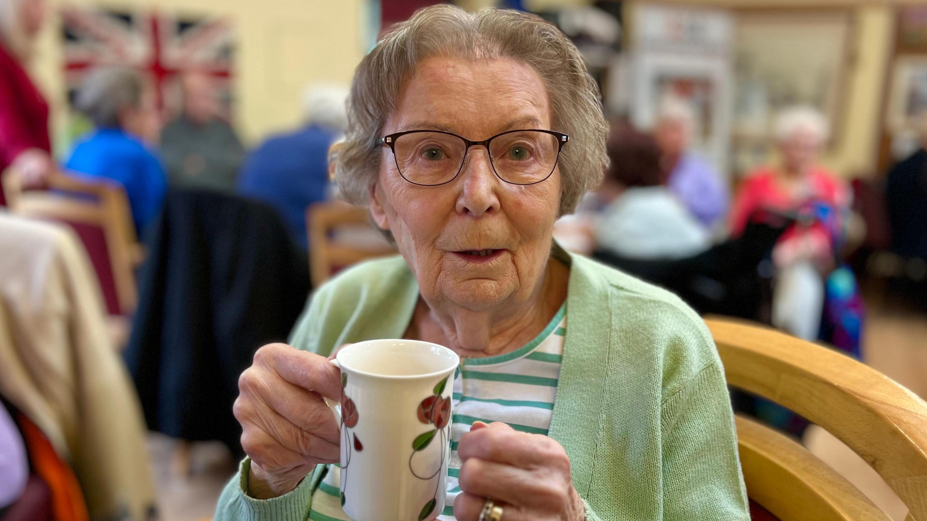 Margaret Rushmore. A lady who is holding a ceramic mug and is looking at the camera. She is wearing a green cardigan and a white and green stripey top. She is also wearing glasses.