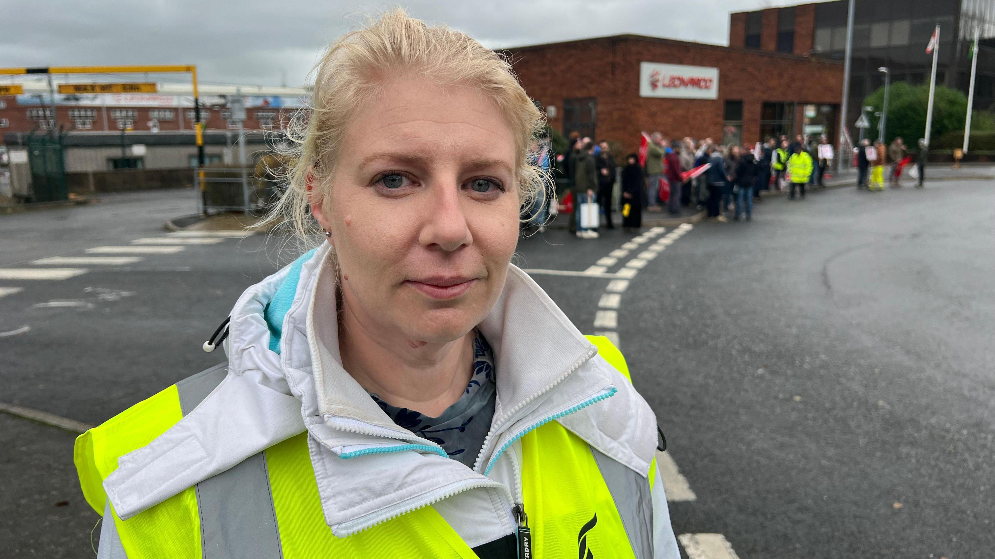 The South West Regional Officer for Unite, Shevaun Hunt wearing a high vis vest and white coat facing the camera. She has blonde hair tied back and is standing outside the front gates of the Yeovil factory.