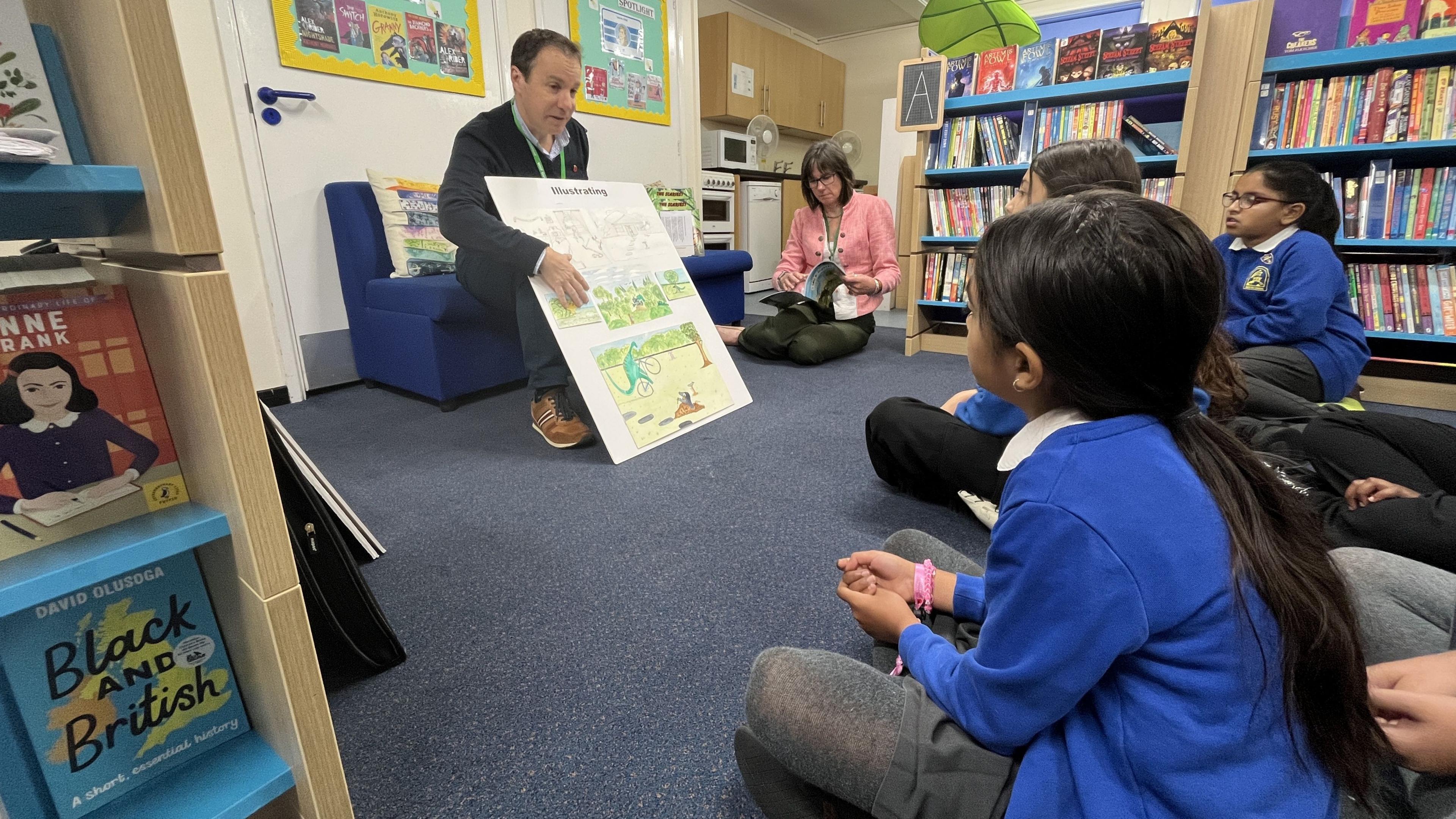 Children sat in a school library. They are crossed legged and looking at a man reading a story board.