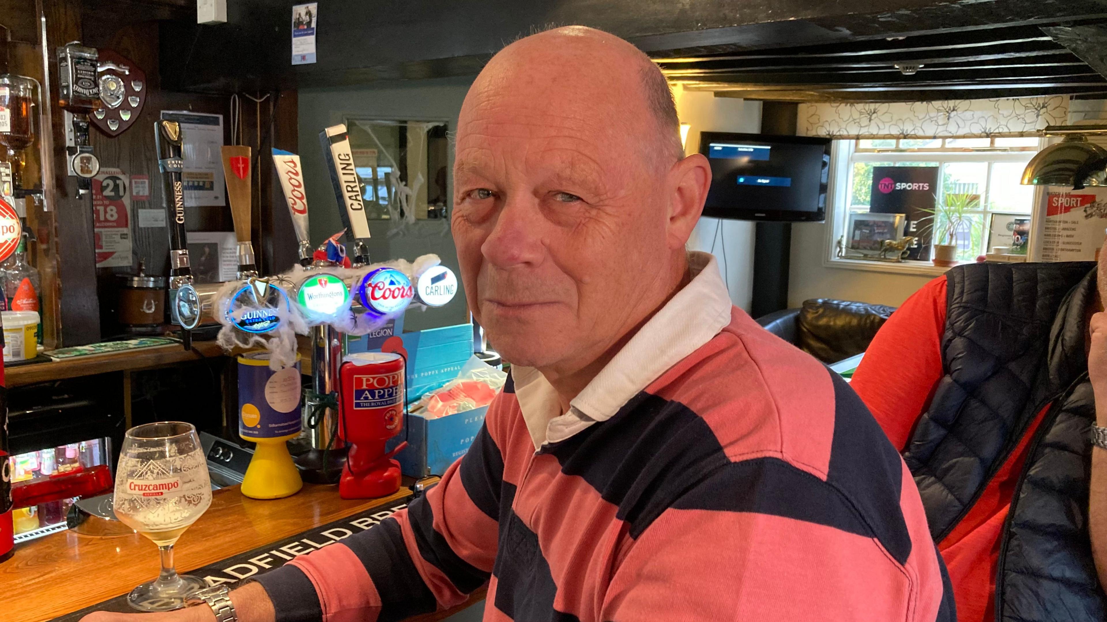 Mike Batt with short hair sitting at a bar in a public house wearing a striped red and black top looking towards the camera. 