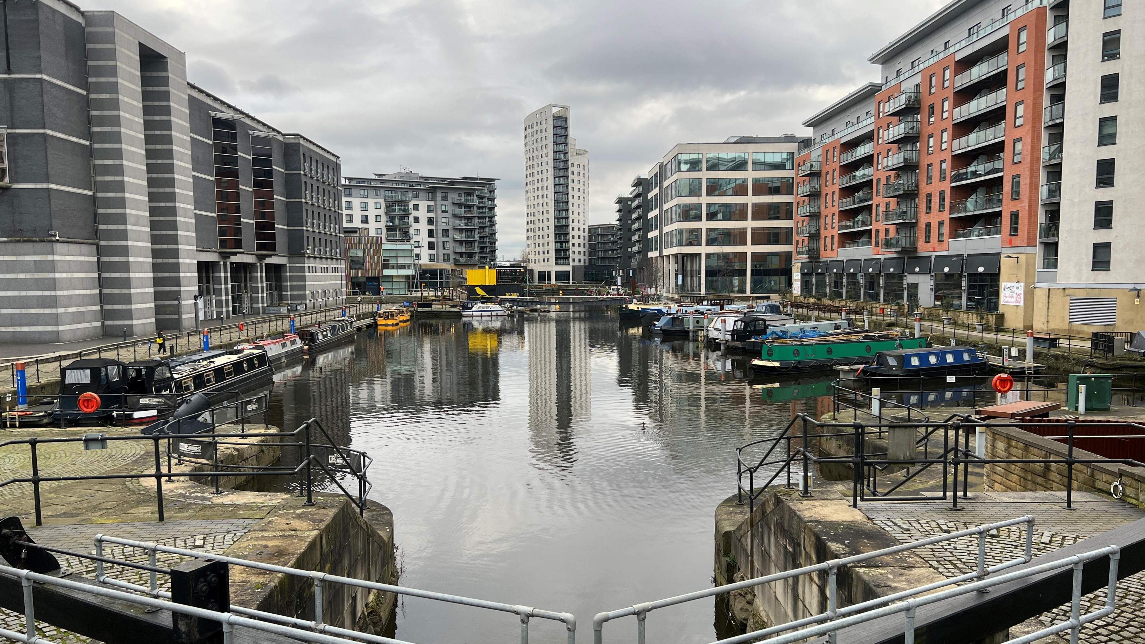 A picture of Leeds Dock, there is water with narrow boats on either side, and buildings surrounding it.