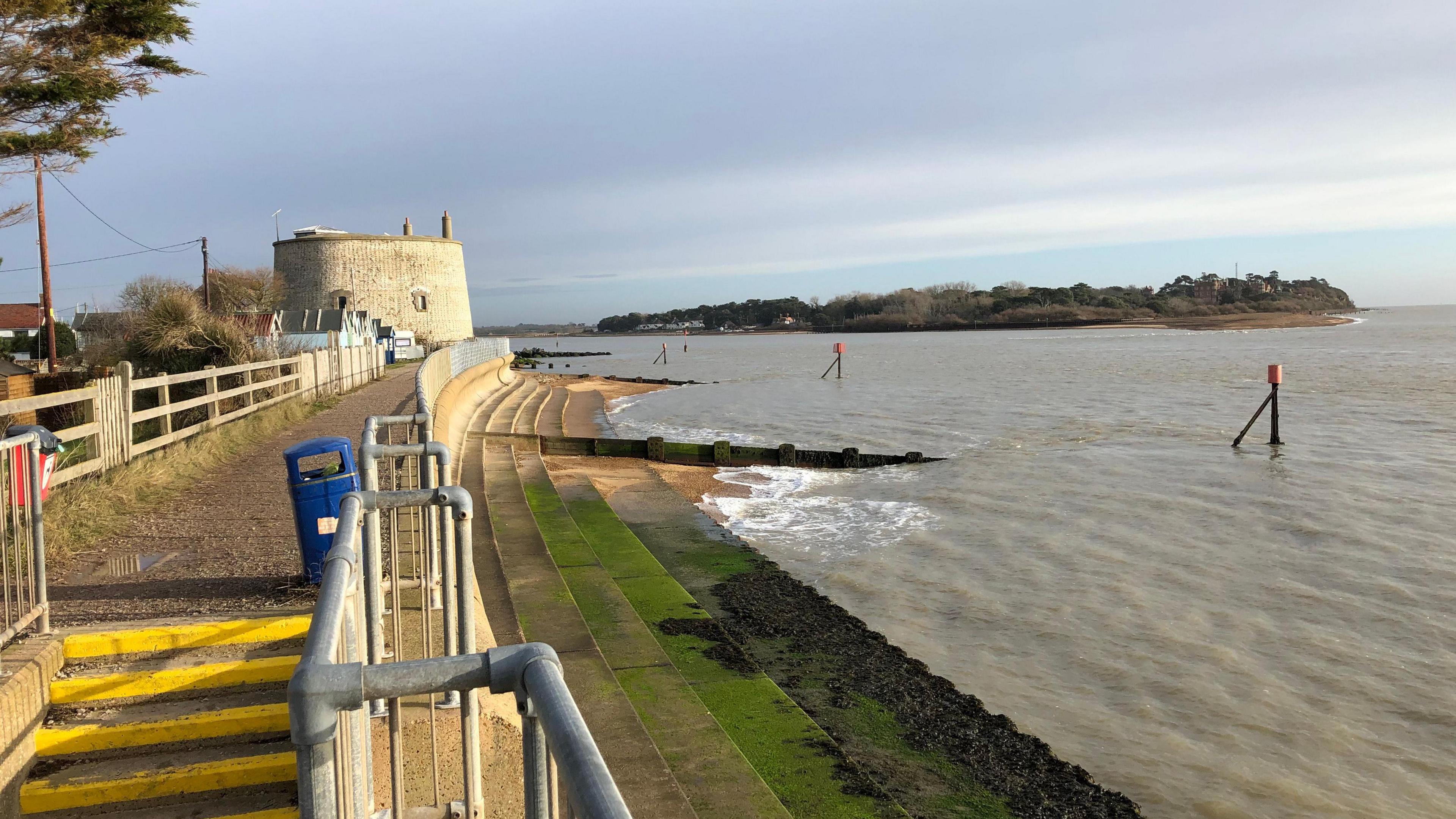 The mouth of the River Deben near Felixstowe. It is high tide and steps leading down on to the beach have been covered by the water. A path along the coast can still be seen above the steps and water.