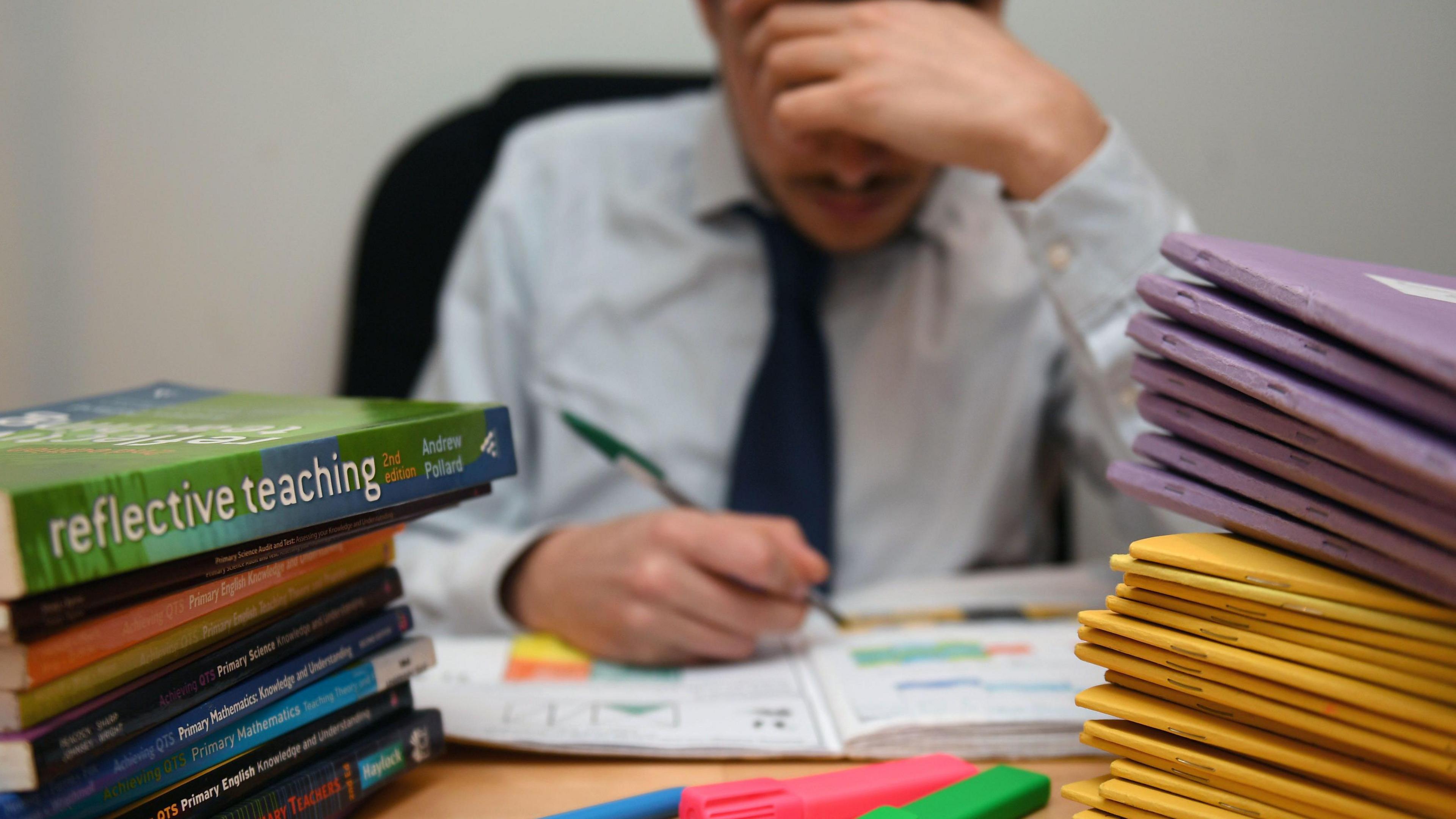 Image of a male teacher working at a desk. His face is shielded by his hand but there are books with titles such as "reflective teaching" in the foreground.