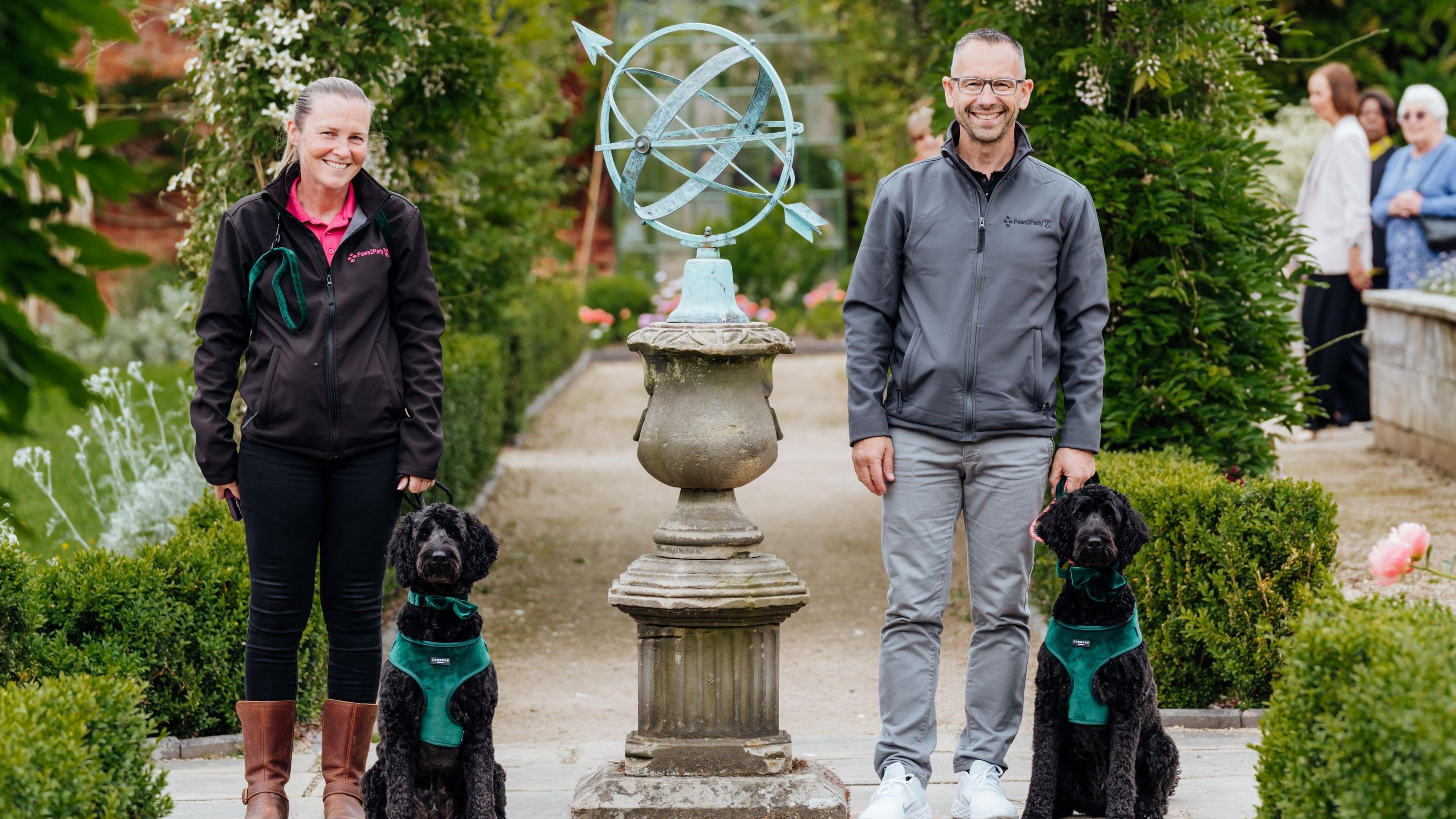 A woman wearing a black jacket and jeans with brown boots, next to a black haired dog with a dark green suede bowtie and harness. Next to her, a man with glasses wearing a grey jacket and jeans with another black haired dog wearing a dark green suede bowtie and harness. 