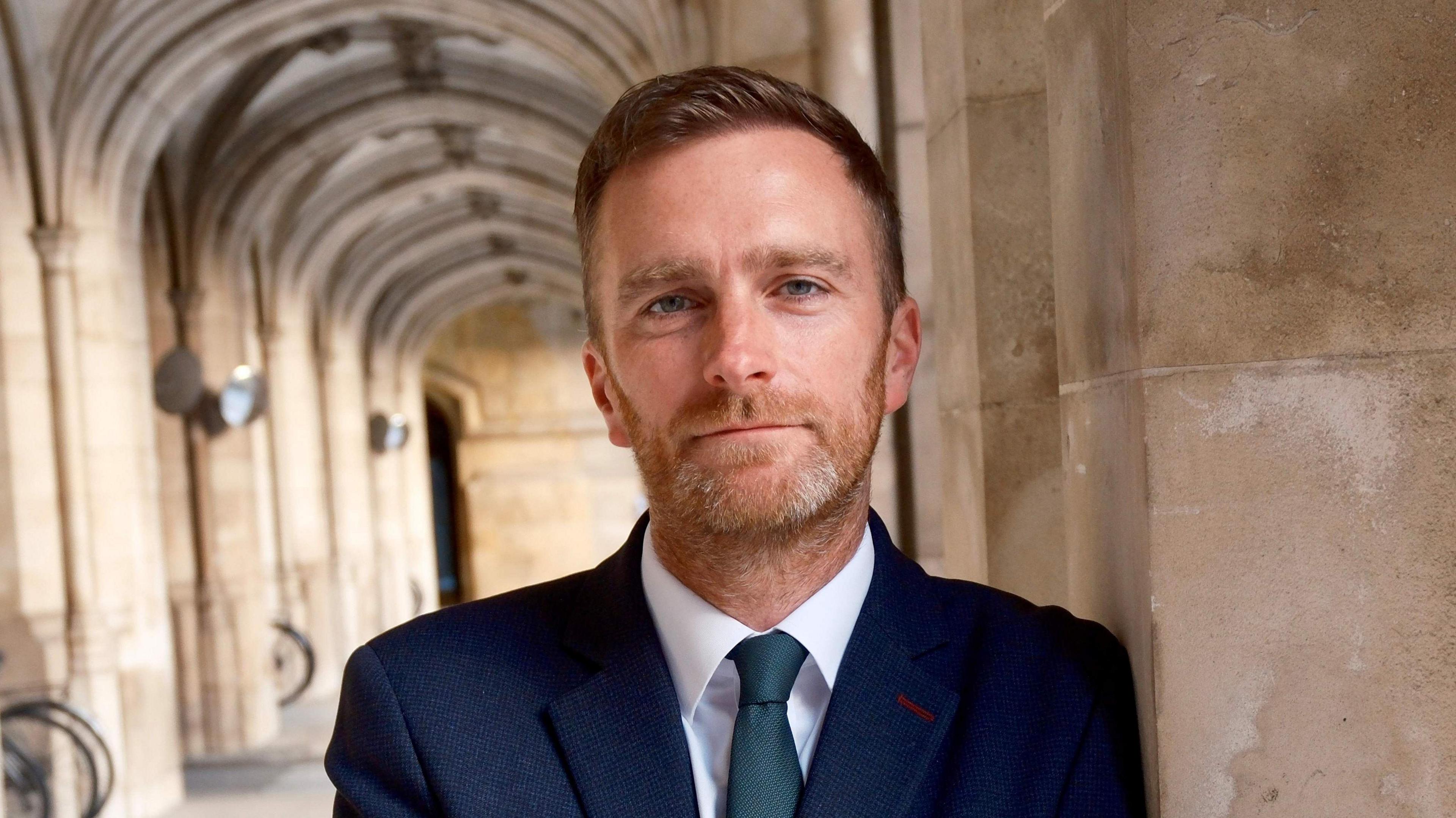 Matt Vickers is dressed in a navy suit and tie and has a short beard and moustache and short, dark blond hair. He is standing in Westminster leaning against a stone pillar with a long, stone corridor extending behind him.