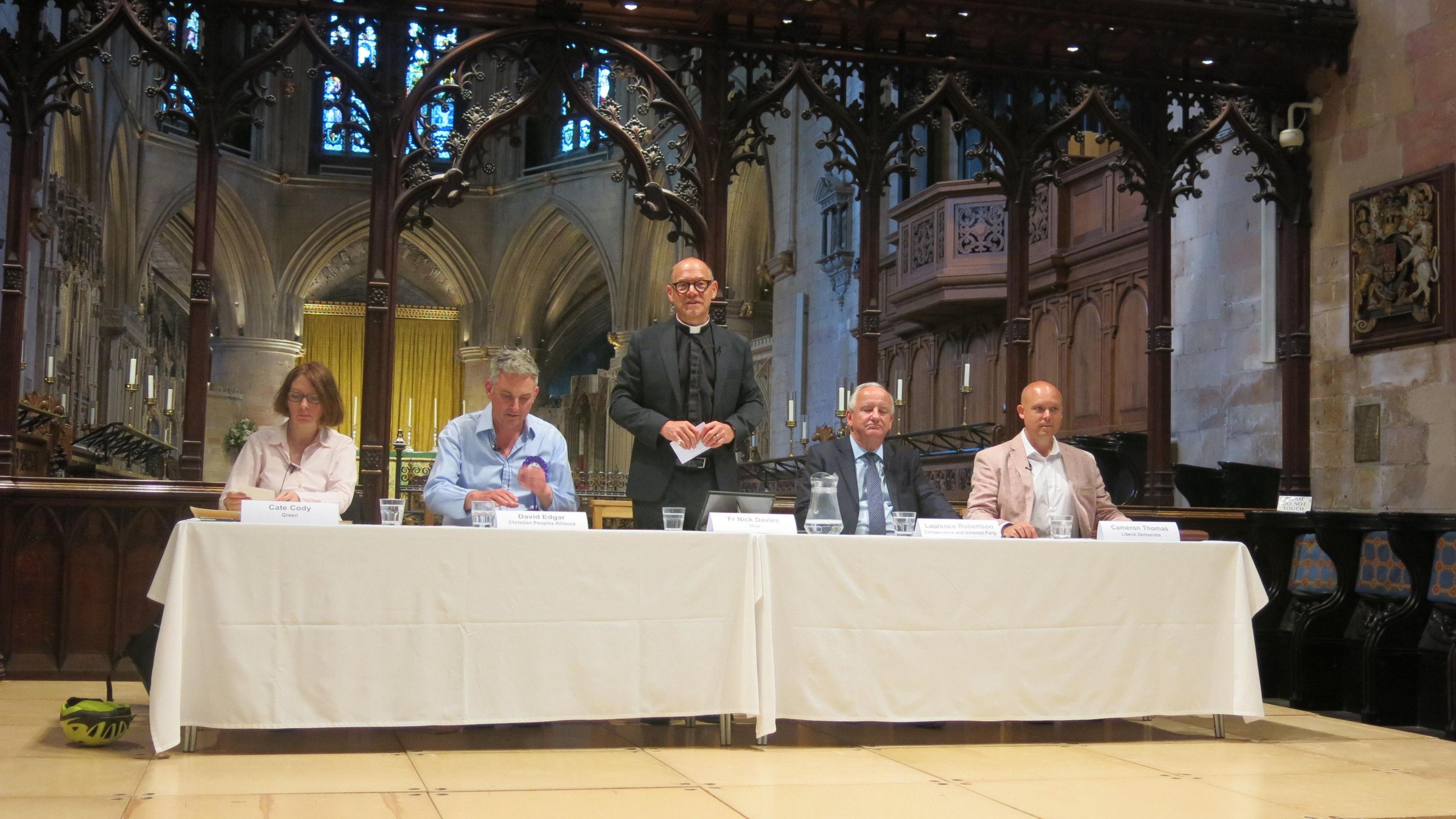 Four people sitting either side of a standing vicar on a stage in an abbey