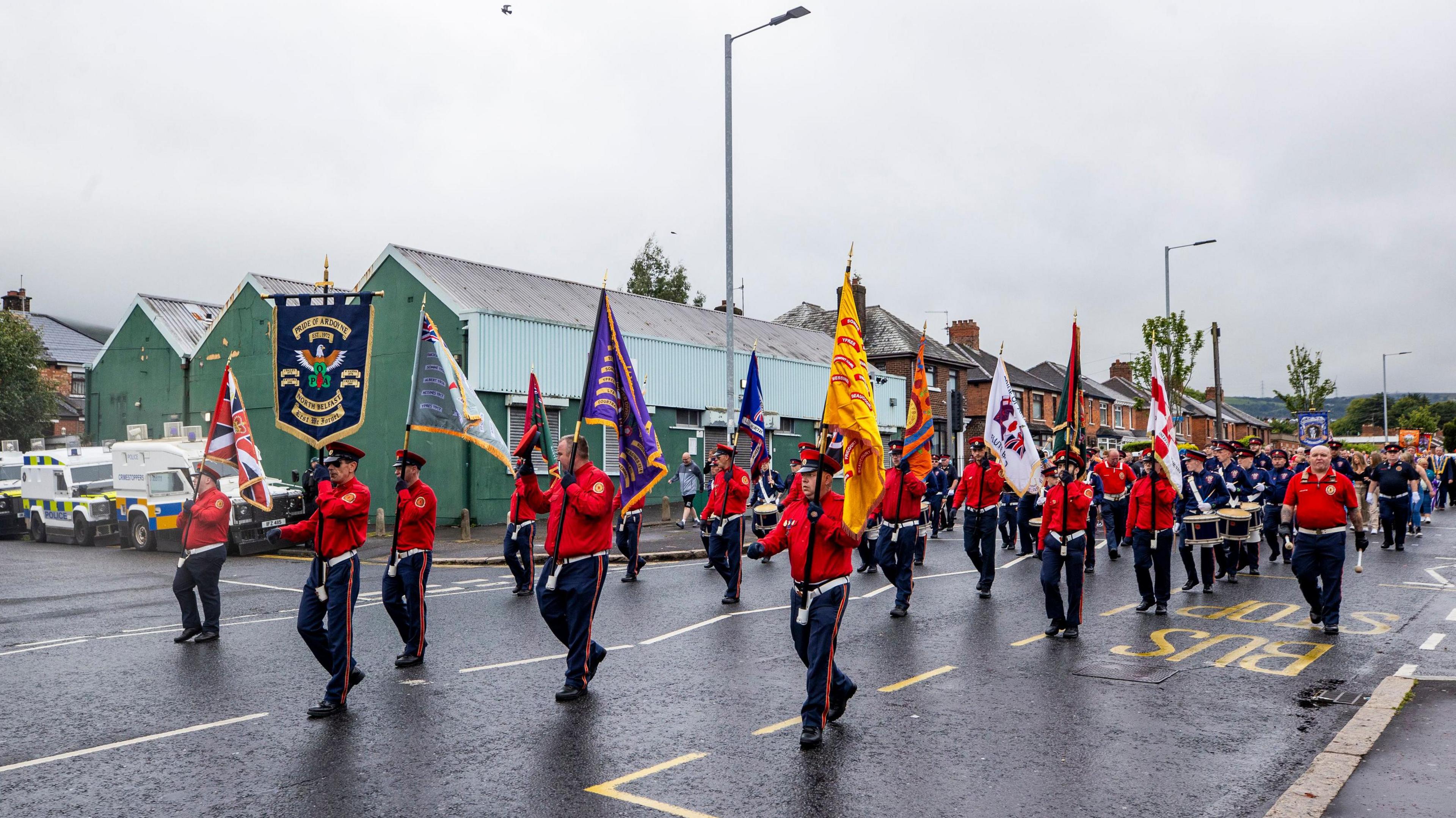 The Pride of Ardoyne flute band led by ranks of men in red shirts and navy trousers holding flats, and behind them ranks of side drummers in navy shirts, and behind them, ranks of flautists. 