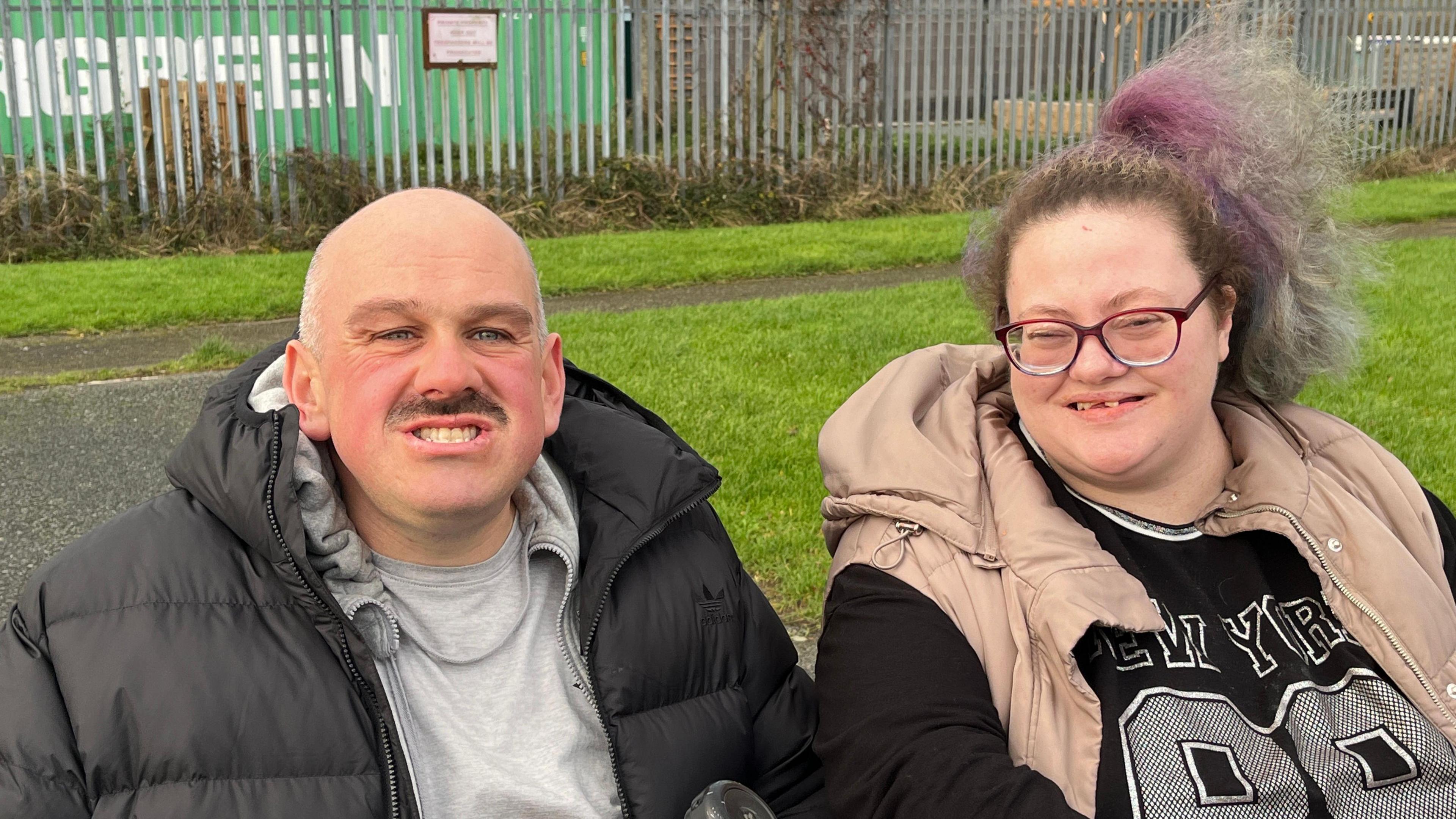 Dyn a menyw gydag anableddau dysgu yn eistedd tu allan ac yn edrych yn syth ymlaen. Mae'r dyn yn foel ac yn gwisgo cot ddu ac mae'r fenyw yn gwisgo sbectol a chot olau heb lewys. Mae ei gwallt hi wedi ei glymu i fyny ac mae'r ddau yn gwenu.