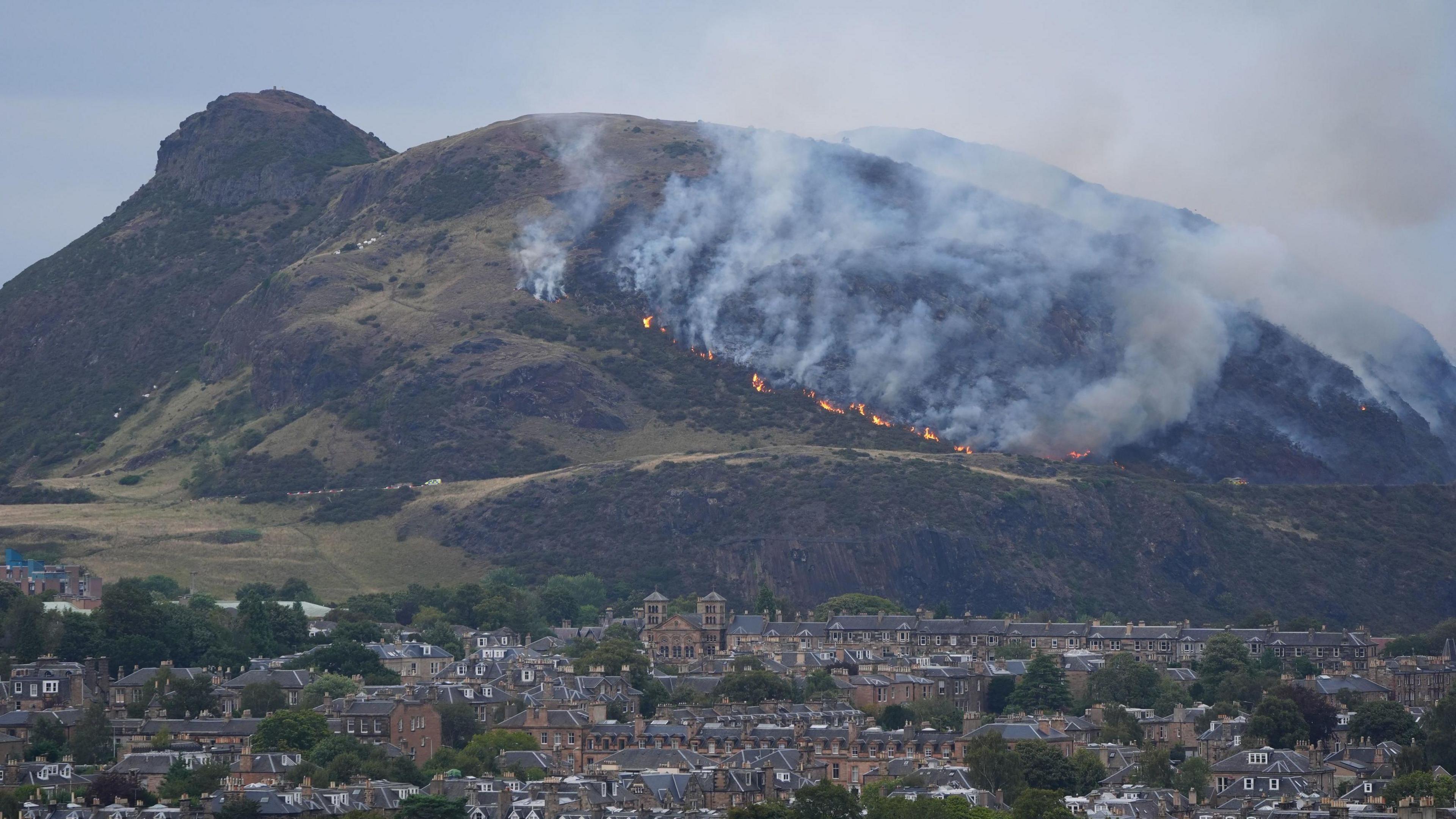 Flames are spreading up a hill. Edinburgh is in the foreground. Smoke can be seen billowing from the flames into the sky