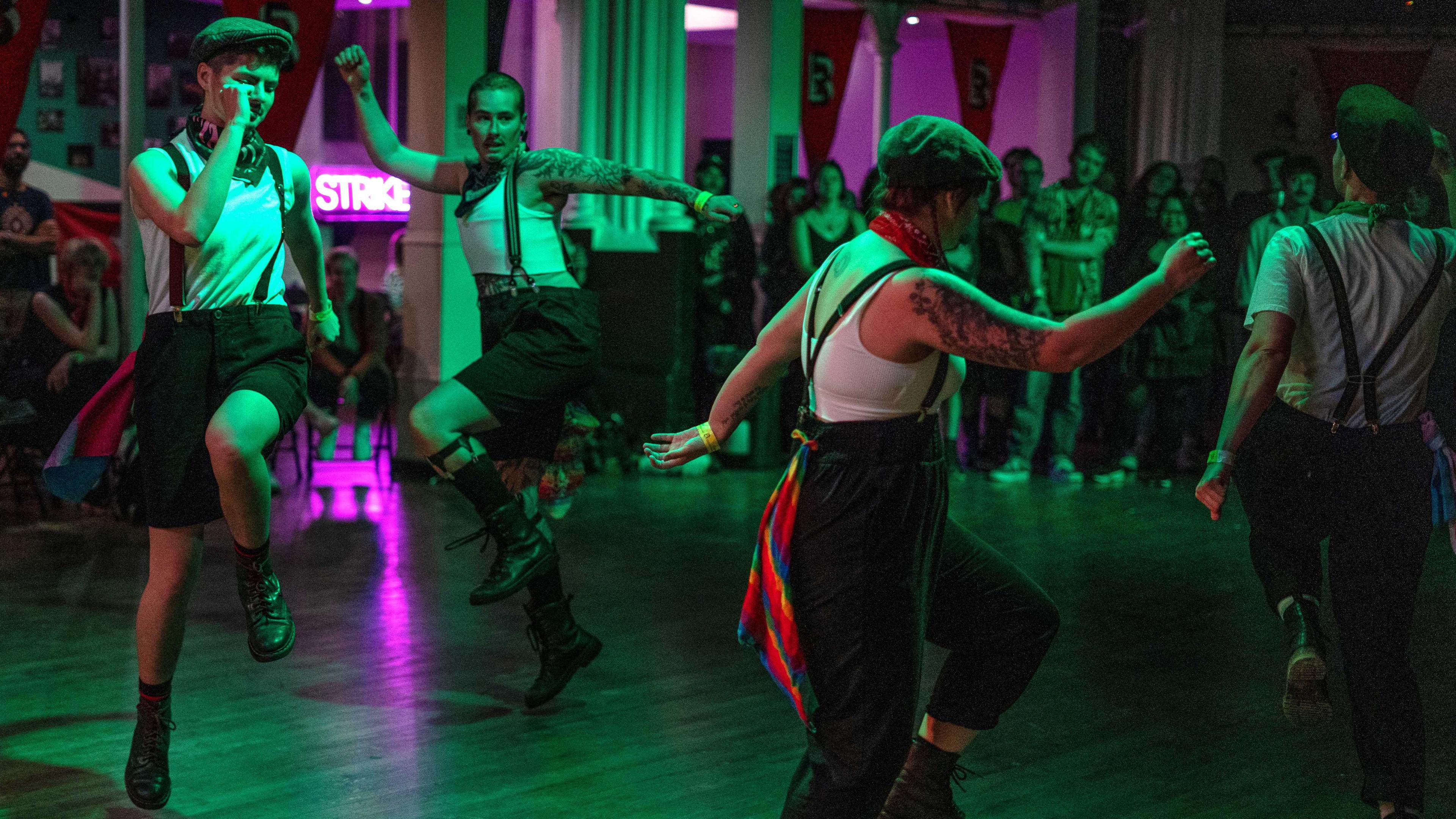 Four dancers dressed in white tops and black shorts or trousers with braces dance in a circle inside a dimly lit room.