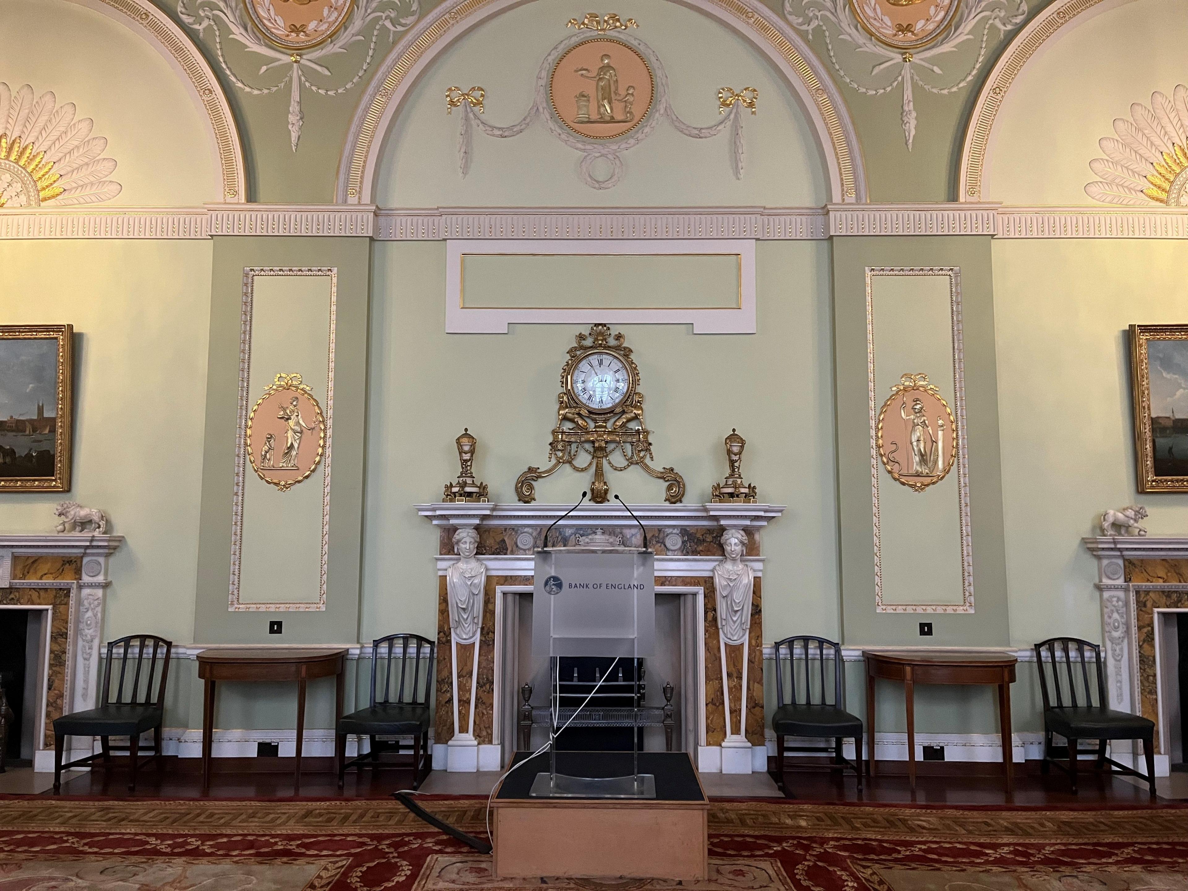 The court room in the Bank of England which has decorated green walls, a gold clock above a fireplace and a patterned rug. A lectern with Bank of England written on it stands in the middle of the room 