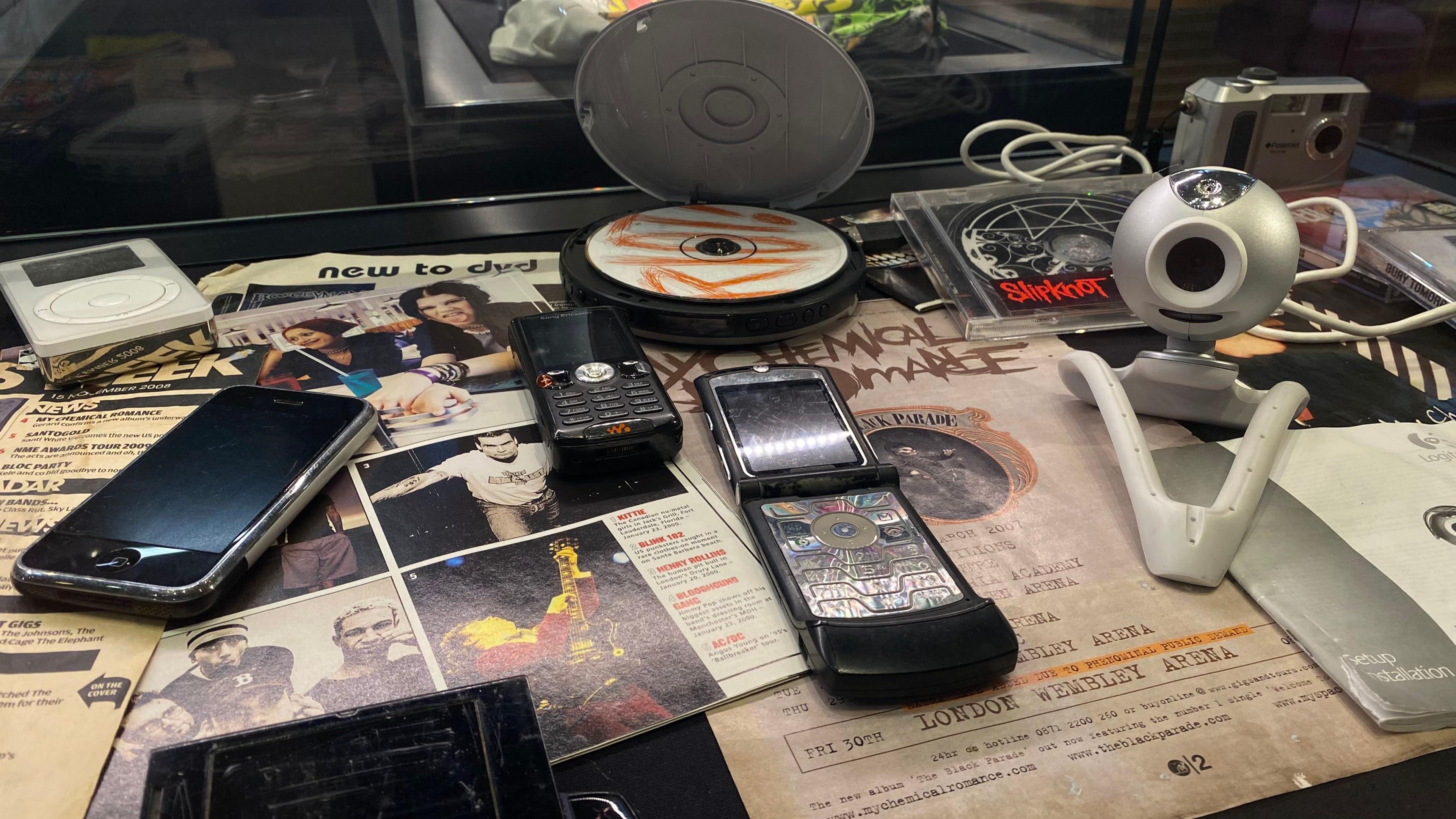 An image of a CD with the words Riot! on. An old iPhone, iPod, a flip phone, webcam and camera. All lay on top of old posters promoting concert dates for popular bands.